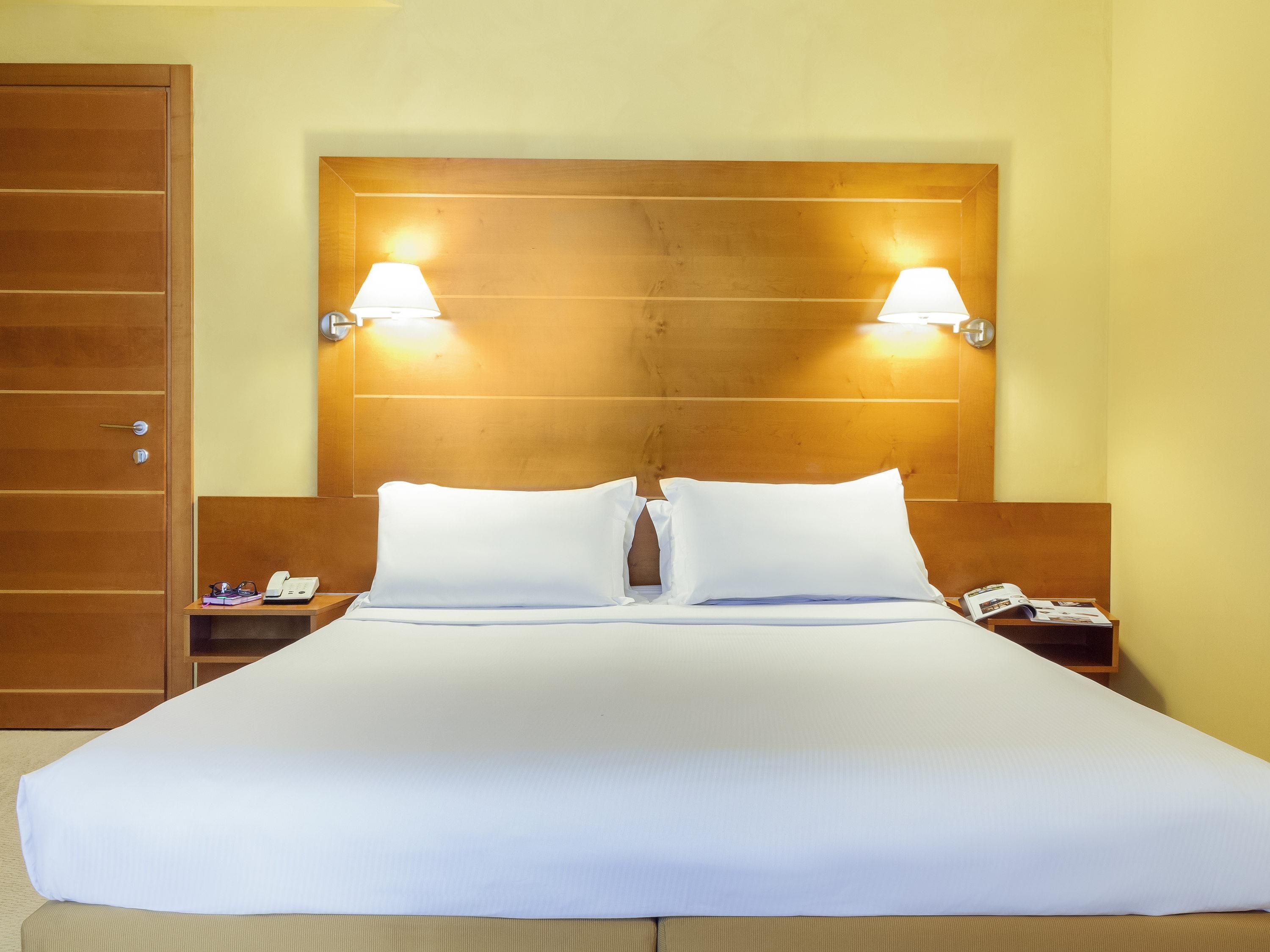 Habitación de hotel con una cama grande, dos mesitas de noche y un cabecero de madera con luces fijadas a la pared.