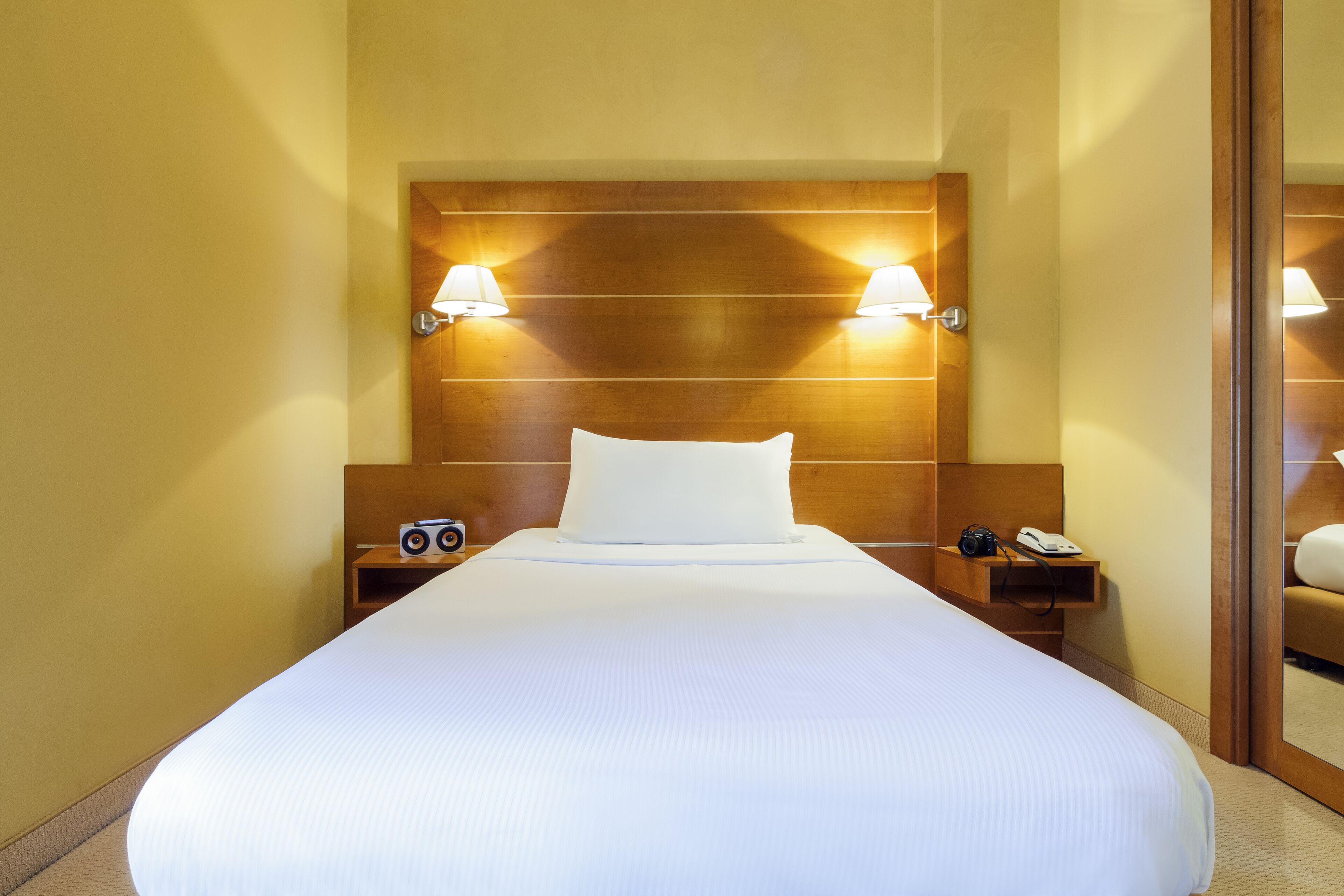 A hotel room with a large bed, wooden headboard, bedside lamps, a nightstand with a camera and phone, and a mirror.
