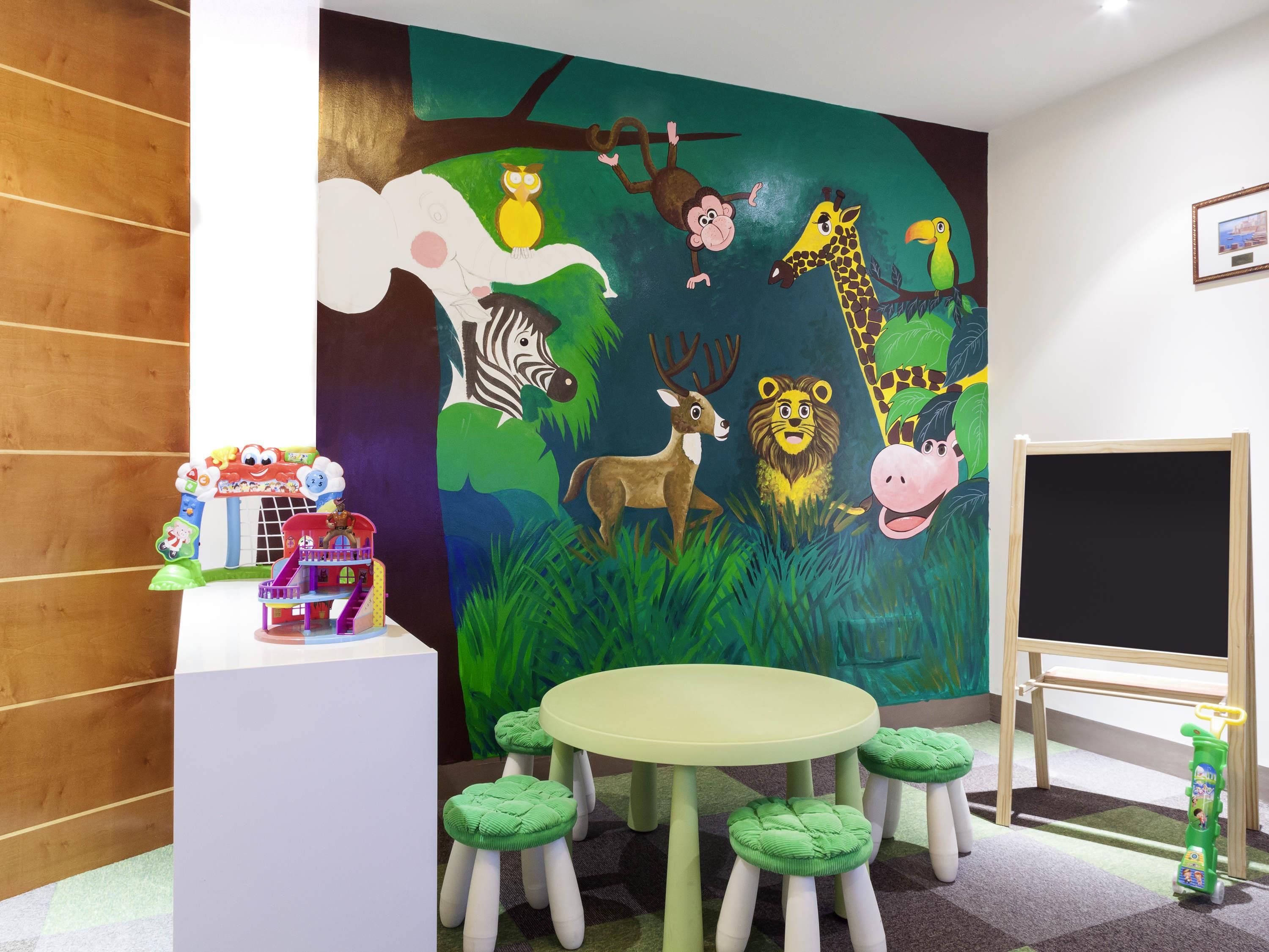 A children's playroom with a mural of animals, a play table with green stools, and a chalkboard.