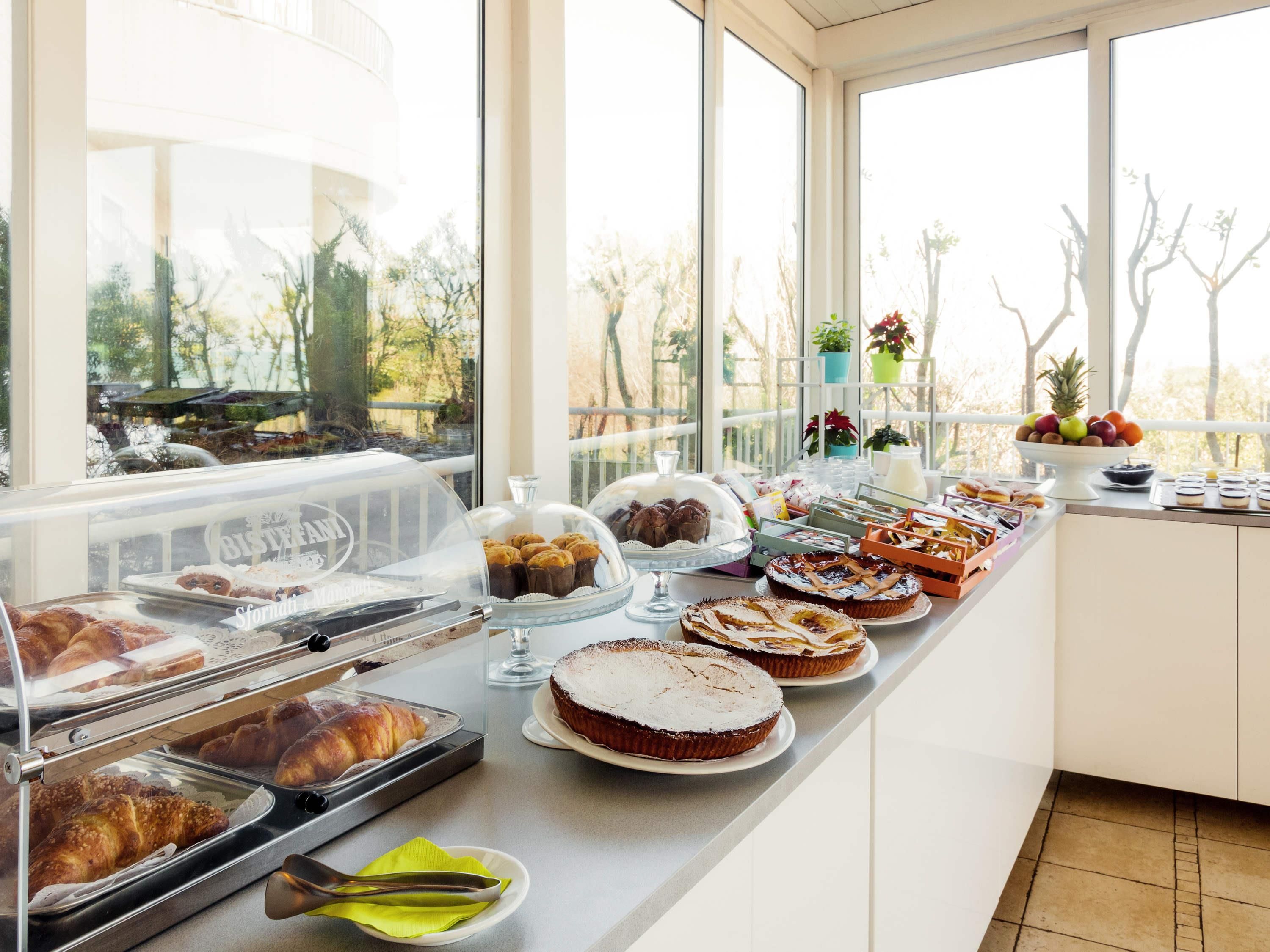En buffet med forskellige bagværk og desserter, et vindue med udsigt til træer, og et lyst, hvidt interiør.