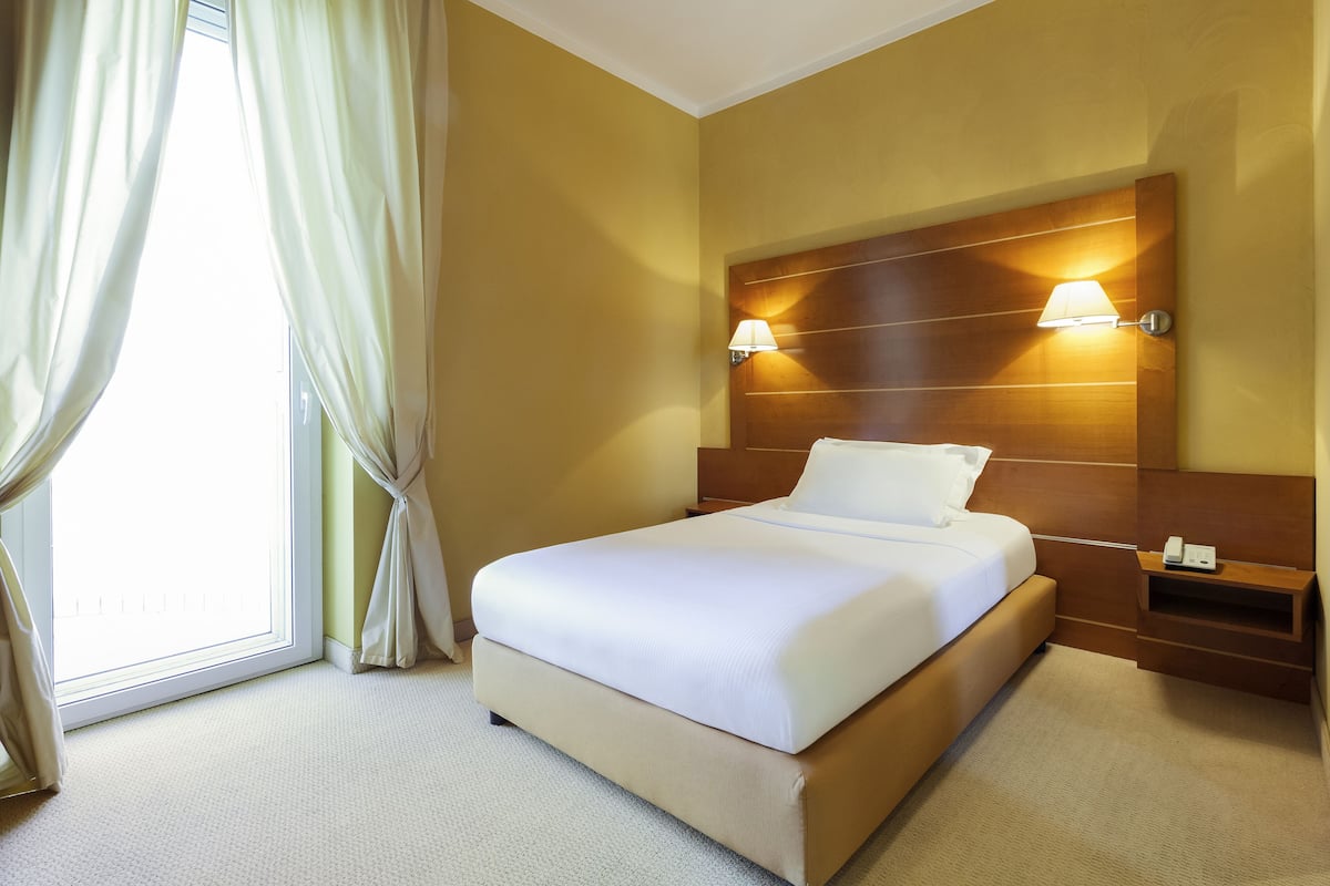 A hotel room with a large bed, a wooden headboard, two bedside lamps, a nightstand, and a window with curtains.