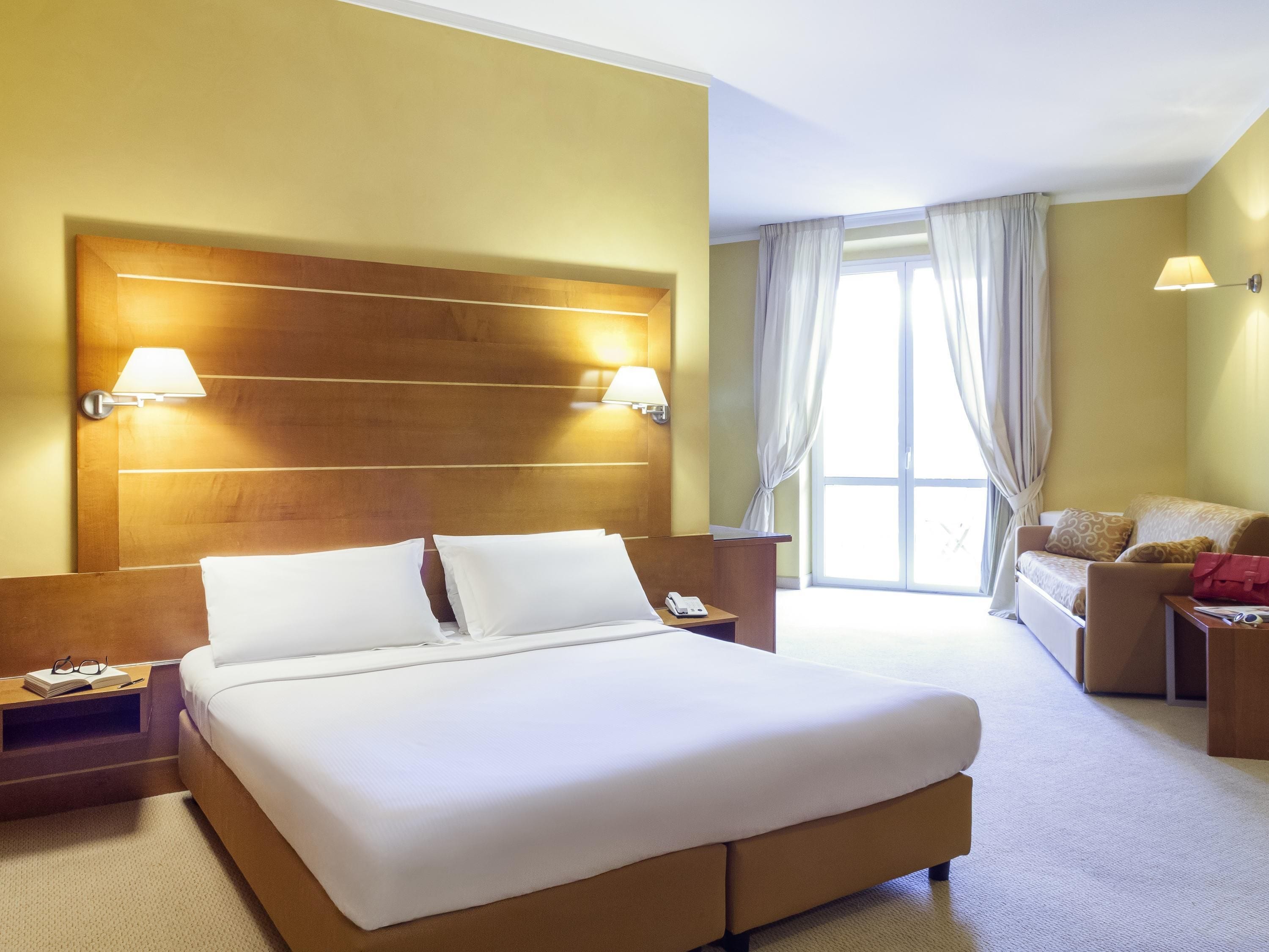 Hotellihuone, jossa on suuri sänky, yöpöydällä lamput, kirjoituspöytä puhelimineen, sohva ja ikkuna, jossa on verhot.