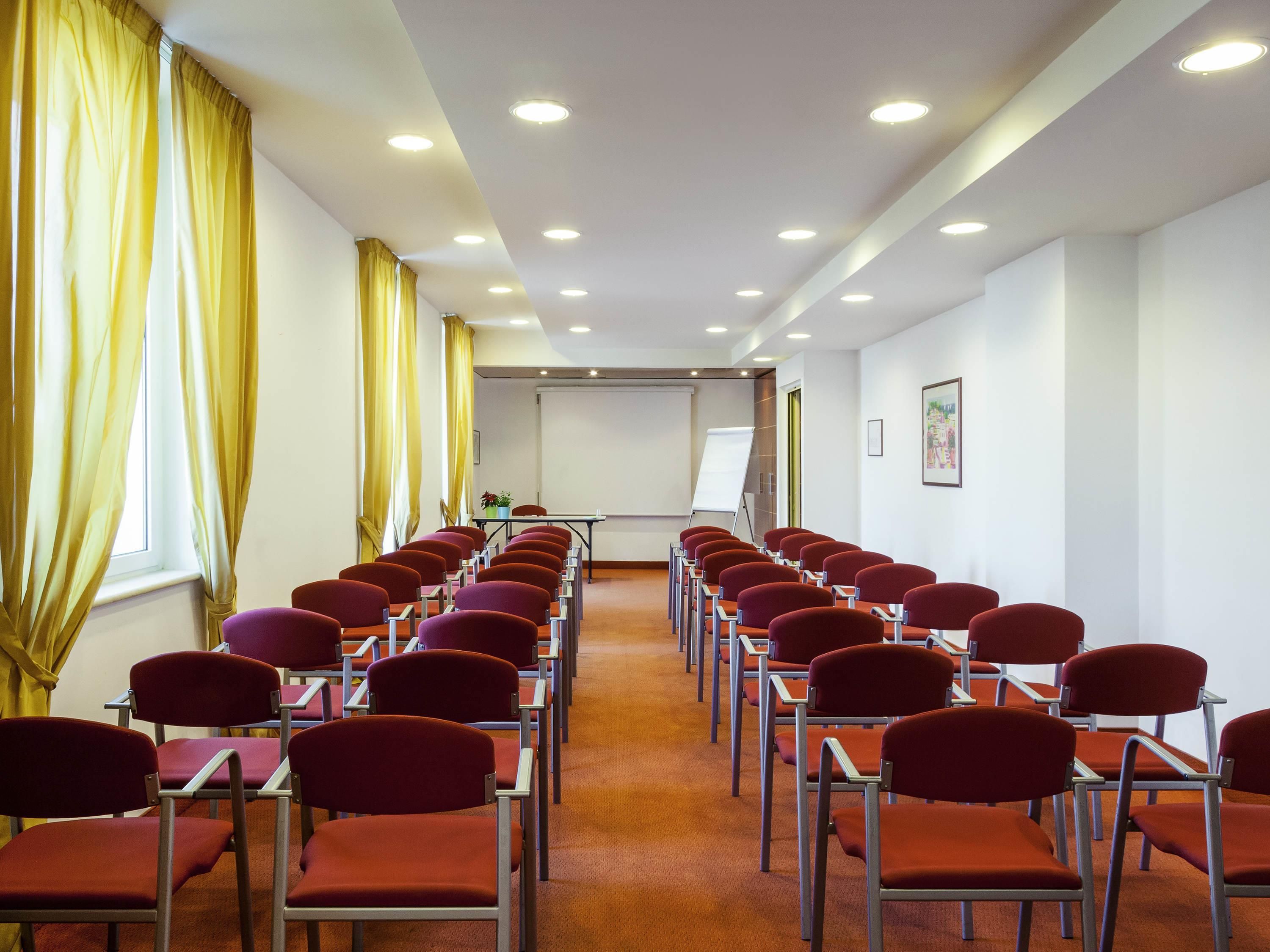 Un salón de conferencias con filas de sillas, una pizarra y una pantalla de proyector.