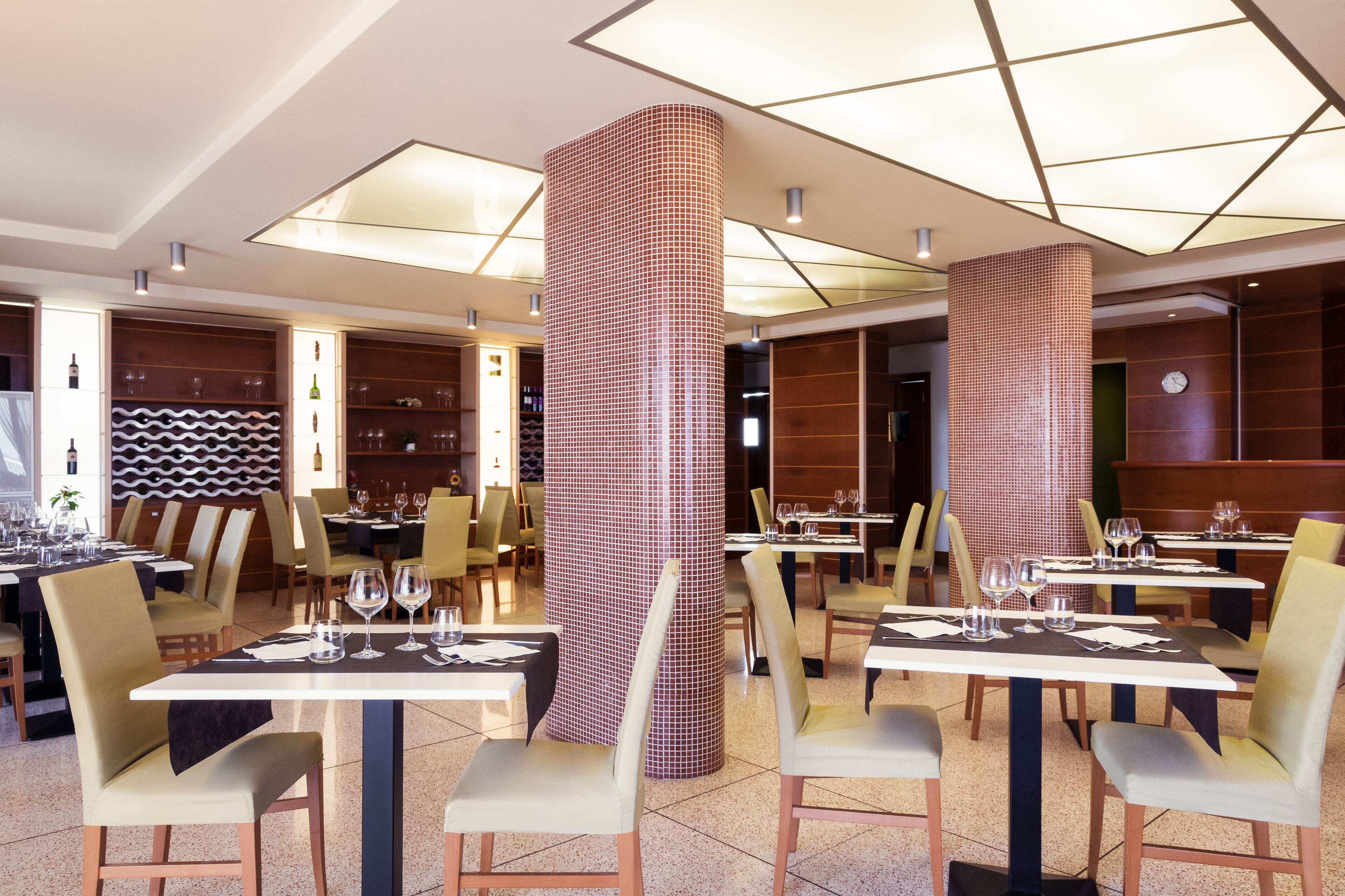 A modern restaurant with high ceilings, geometric ceiling lights, and a central column with a patterned design. Tables are set with white tablecloths and chairs are upholstered in light colors.