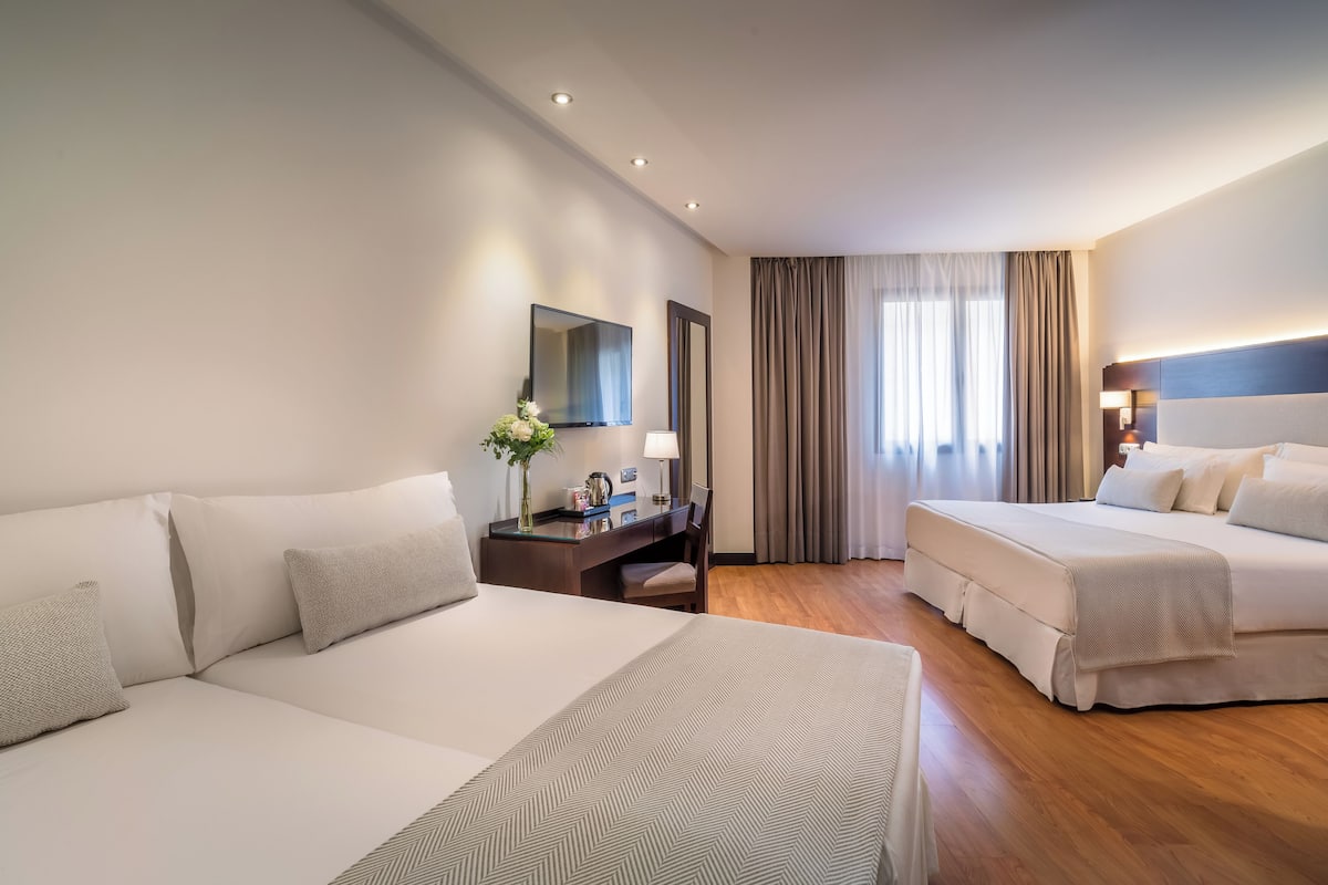 Une chambre d’hôtel avec deux lits, un bureau, une chaise, une télévision et une grande fenêtre avec des rideaux.