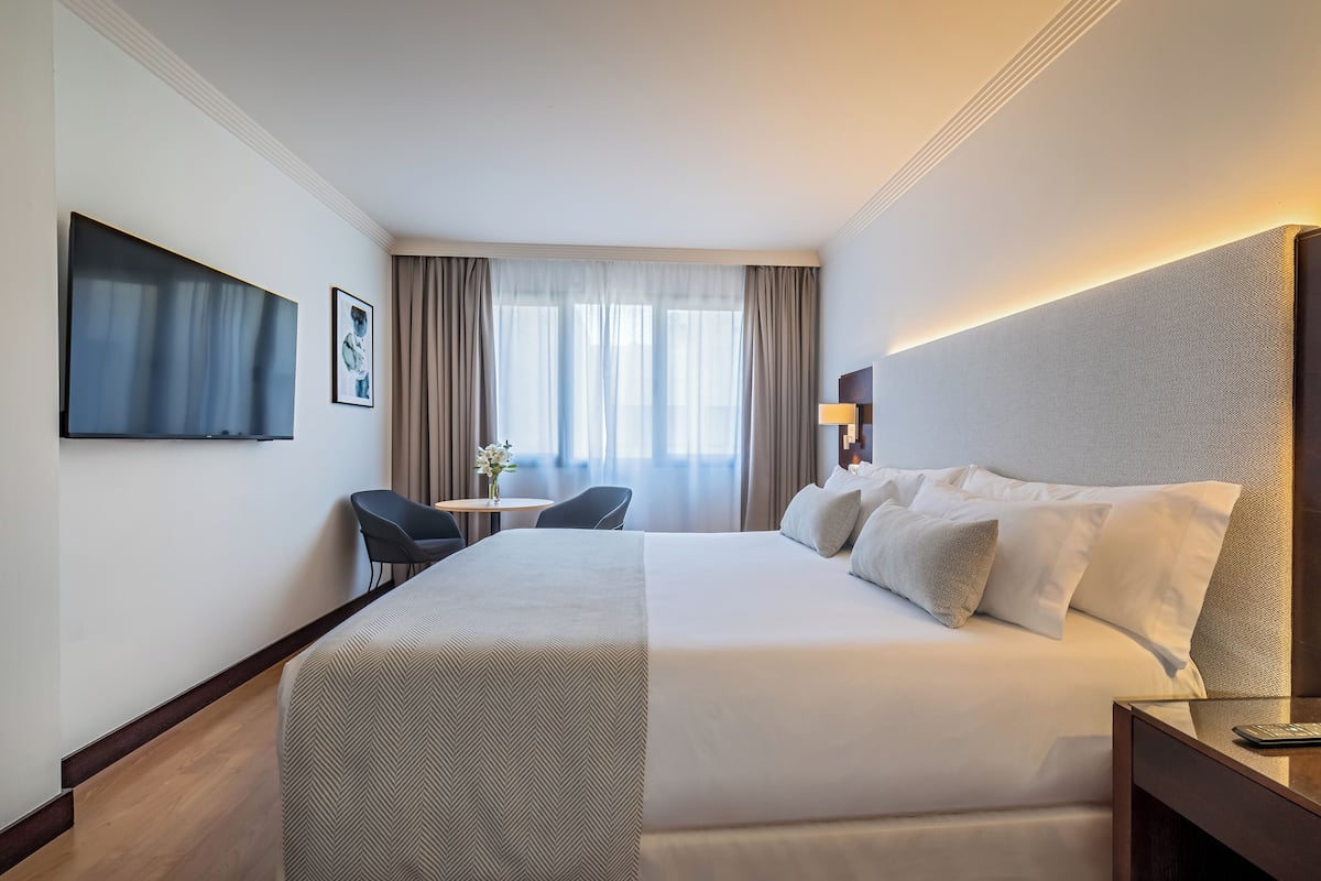 Une chambre d’hôtel avec un grand lit, un bureau, une chaise, une télévision et une fenêtre avec des rideaux.