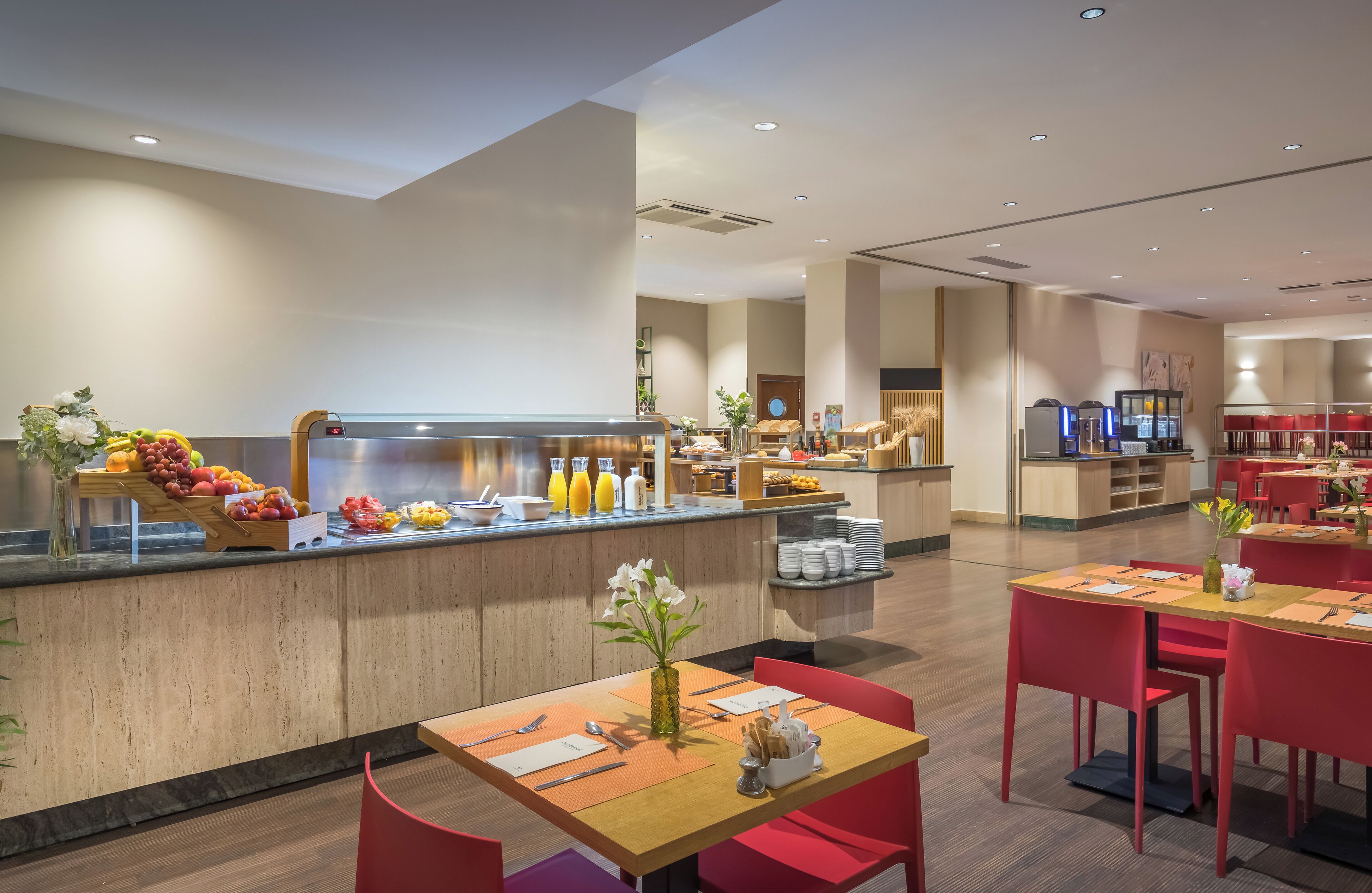 A modern restaurant with a wooden counter displaying fruits and beverages, red chairs, and a clean, minimalist interior.