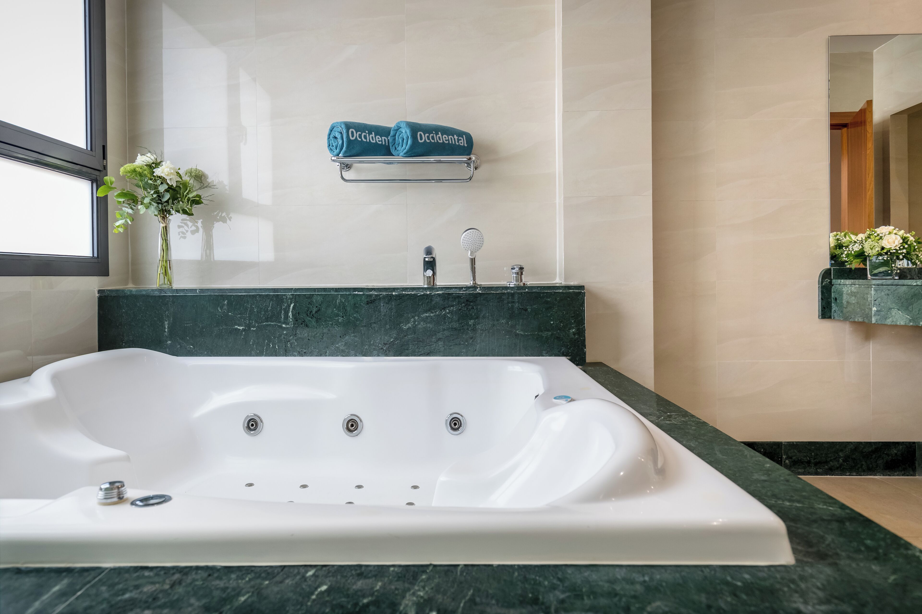 A modern bathroom with a large white bathtub, a marble countertop, a towel rack with folded towels, a vase with flowers, and a mirror.