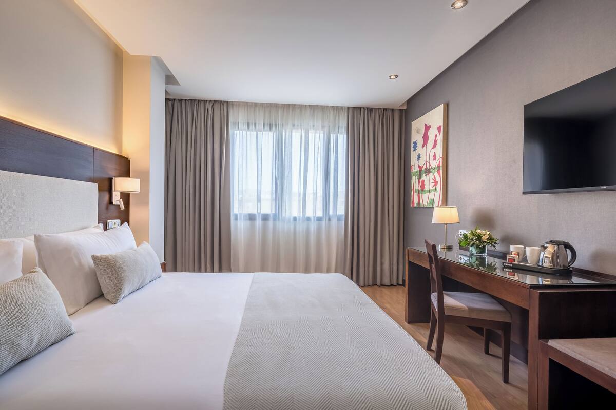 Une chambre d’hôtel avec un grand lit, un bureau, une chaise, un téléviseur à écran plat et une fenêtre avec des rideaux.