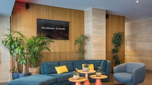 Een moderne lobby met een gebogen blauwe bank, houten paneelwanden en een tv met daarop 'Occidental Granada'.