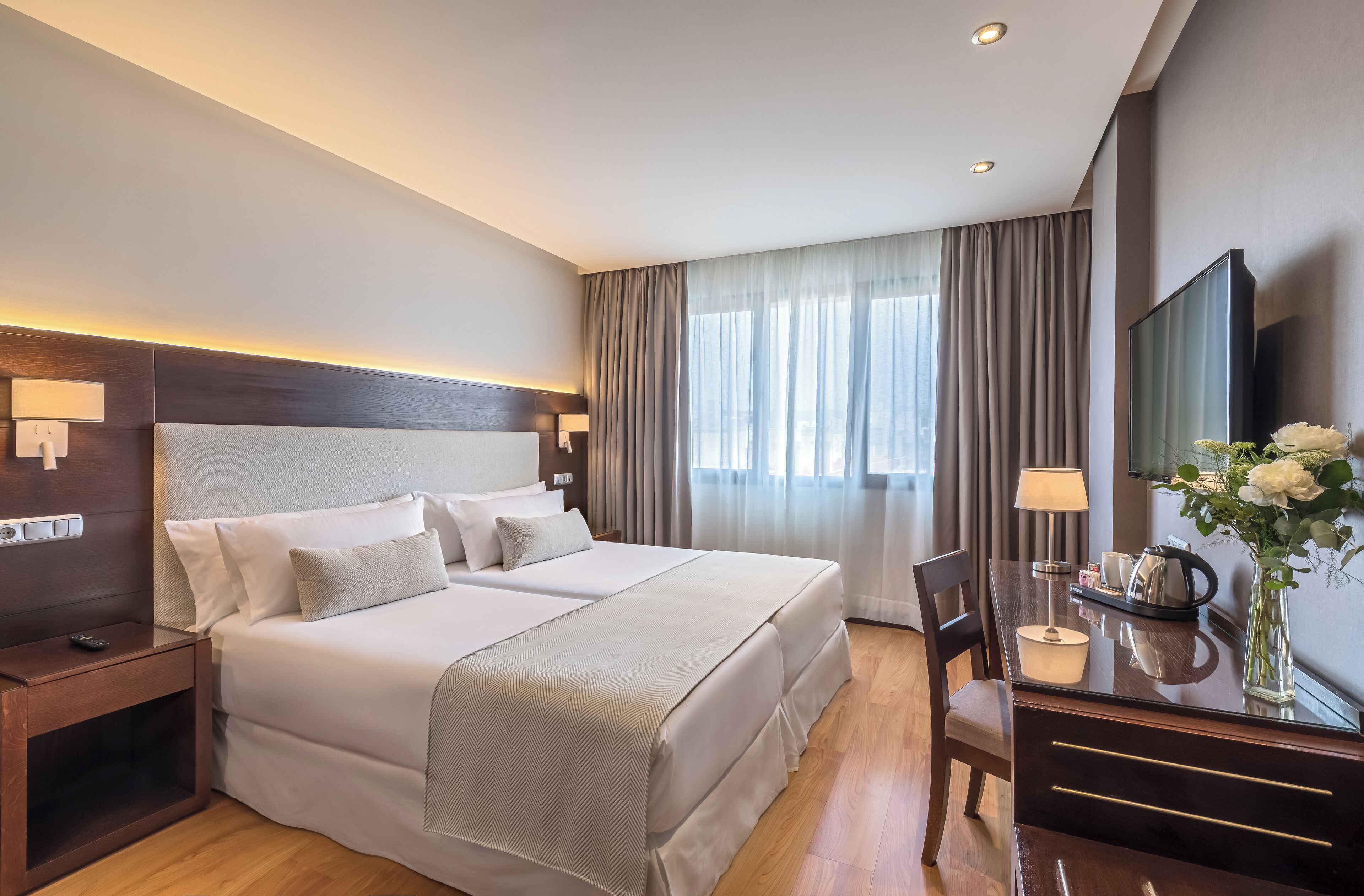 A hotel room with a large bed, bedside tables, a desk with a chair, a TV, and a window with curtains.
