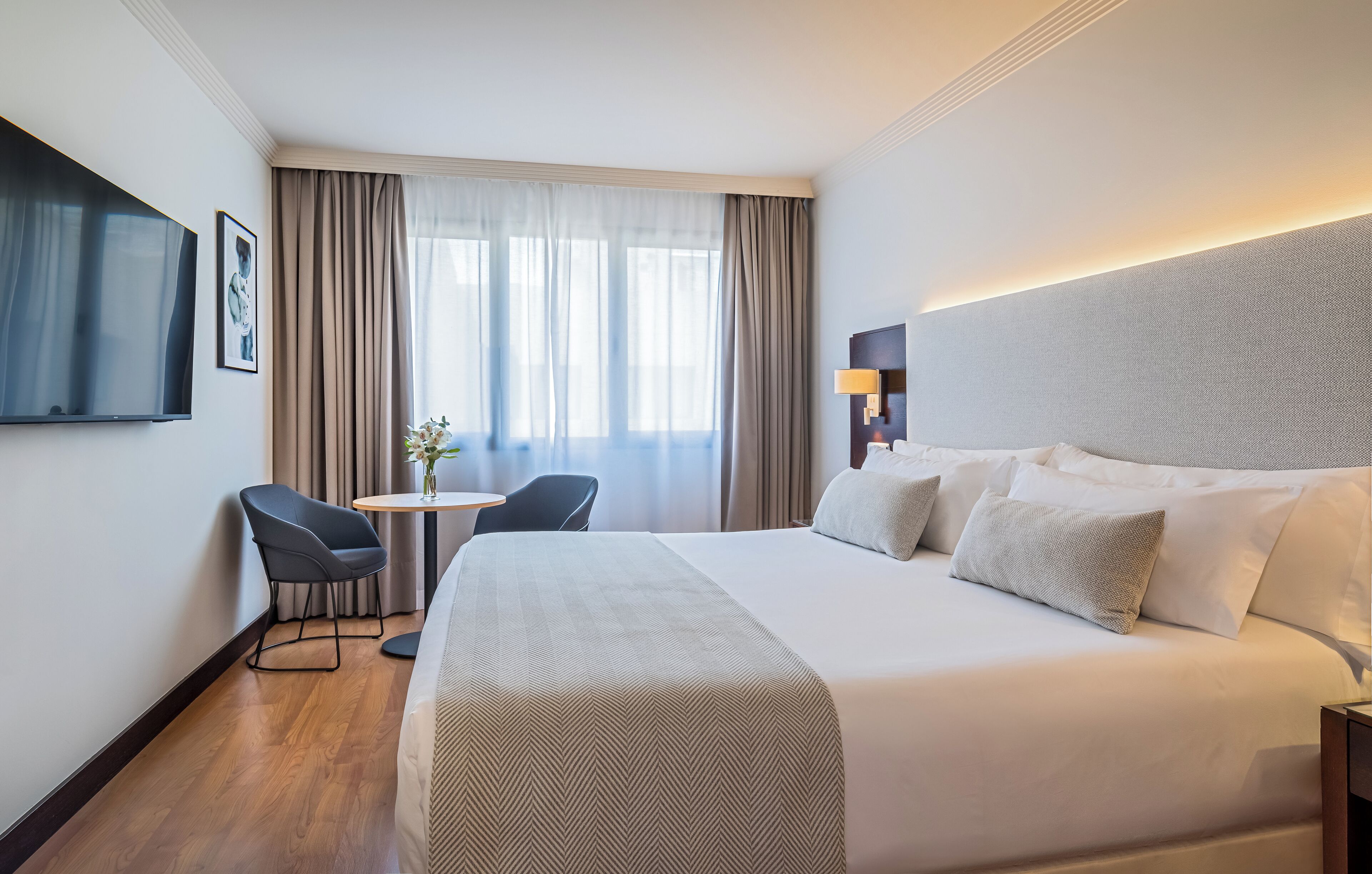 Une chambre d’hôtel avec un grand lit, un téléviseur fixé au mur, un bureau avec une chaise et une fenêtre avec des rideaux.