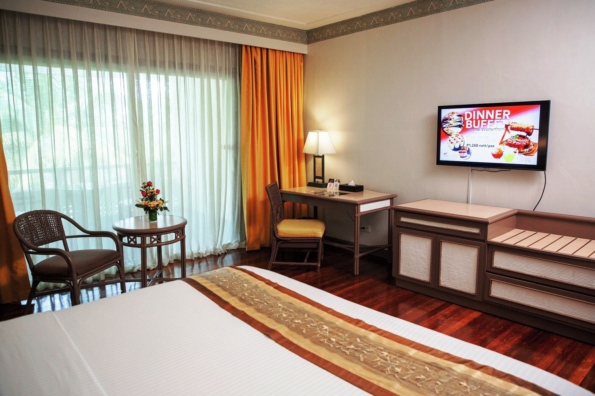 A hotel room with a bed, a desk with a lamp, a TV, and a small table with flowers.