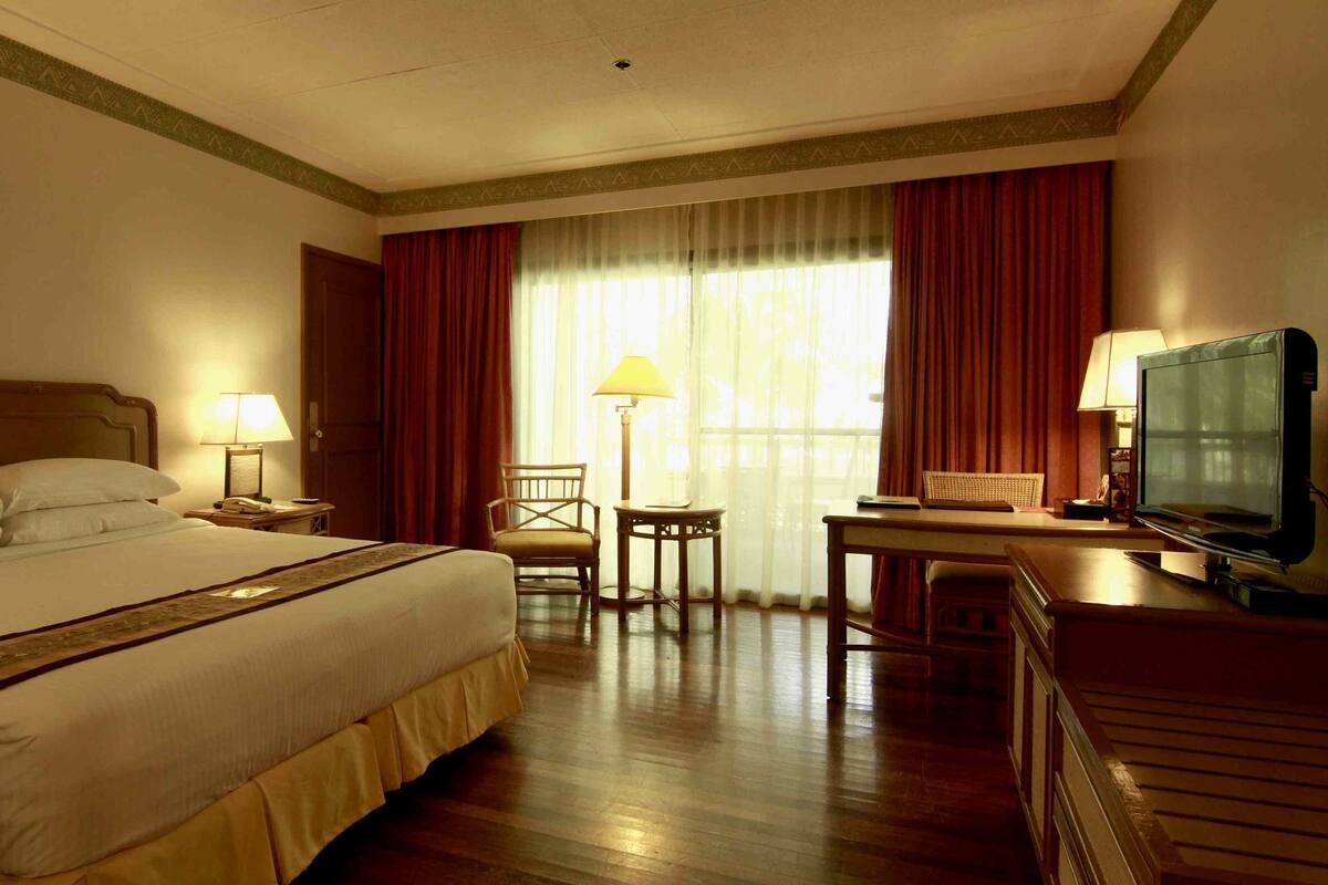 A hotel room with a bed, a desk with a television, a chair, a lamp, and a window with curtains.