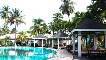 Un área de piscina con sillones de descanso, palmeras y zonas de estar cubiertas.