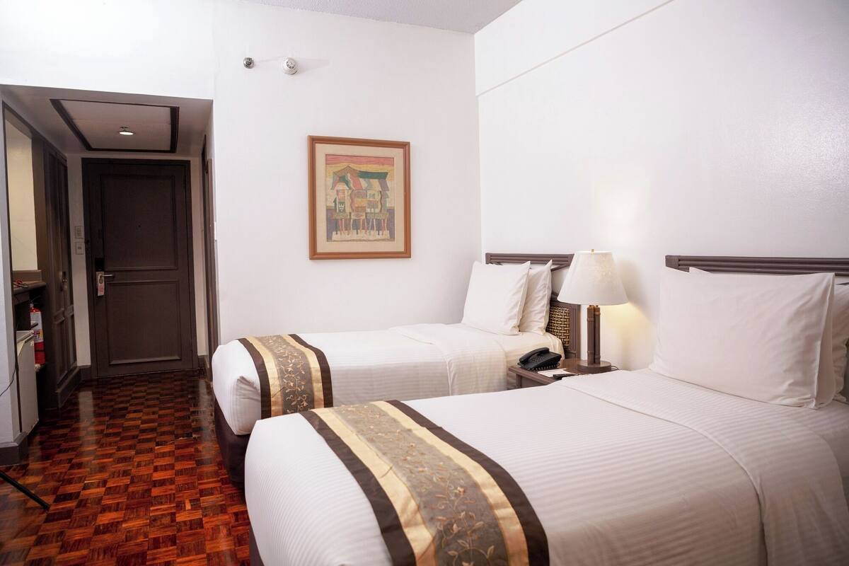 A hotel room with two beds, a wooden floor, a painting on the wall, and a nightstand with a lamp.