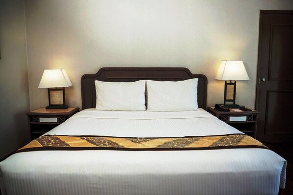 Una cama bien hecha con una colcha blanca y dos almohadas, flanqueada por dos mesitas de noche con lámparas, en una habitación con cabecera de madera y una puerta cerrada.