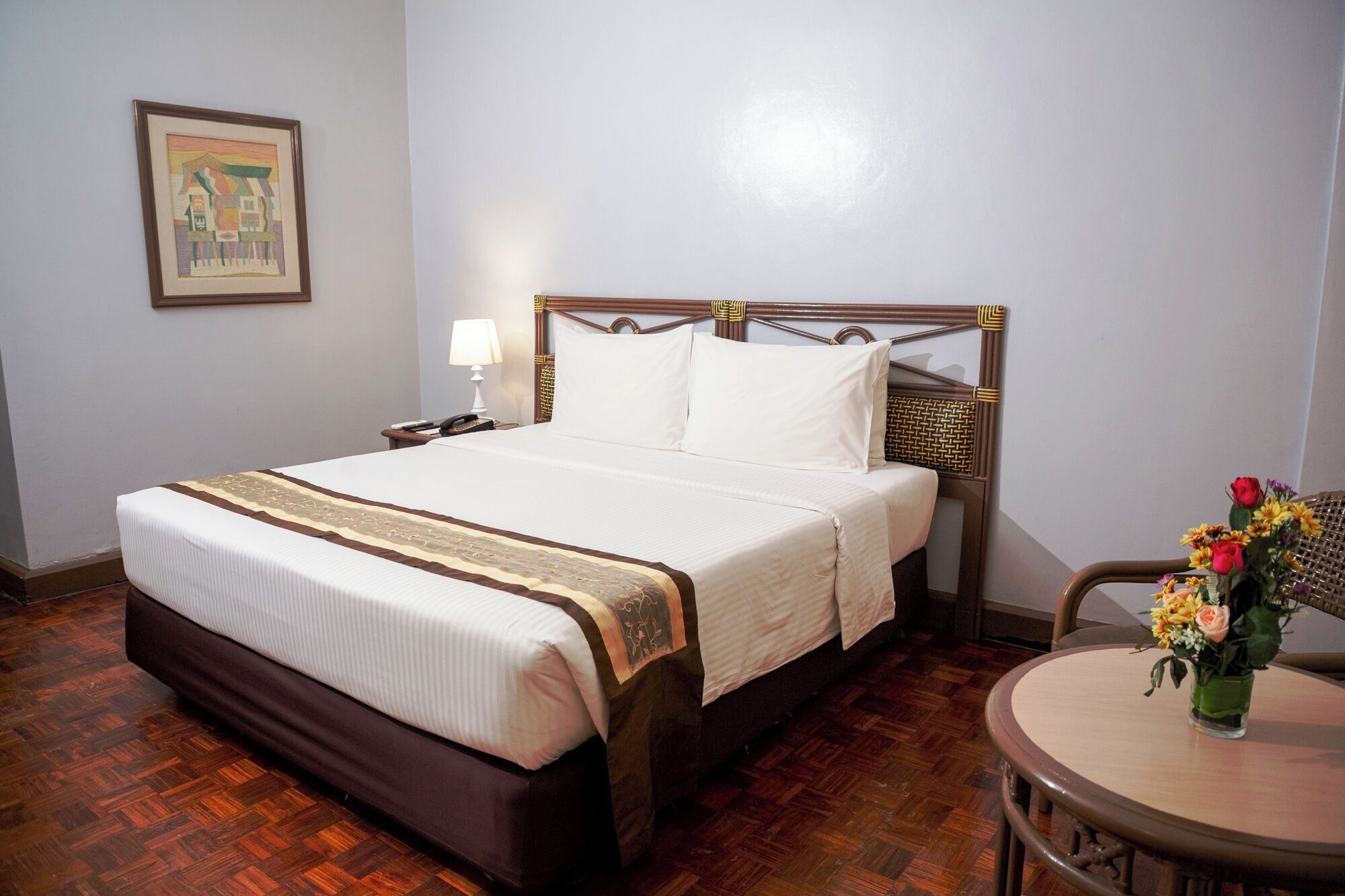 A hotel room with a large bed, a bedside table with a lamp, a chair, and a small table with flowers.