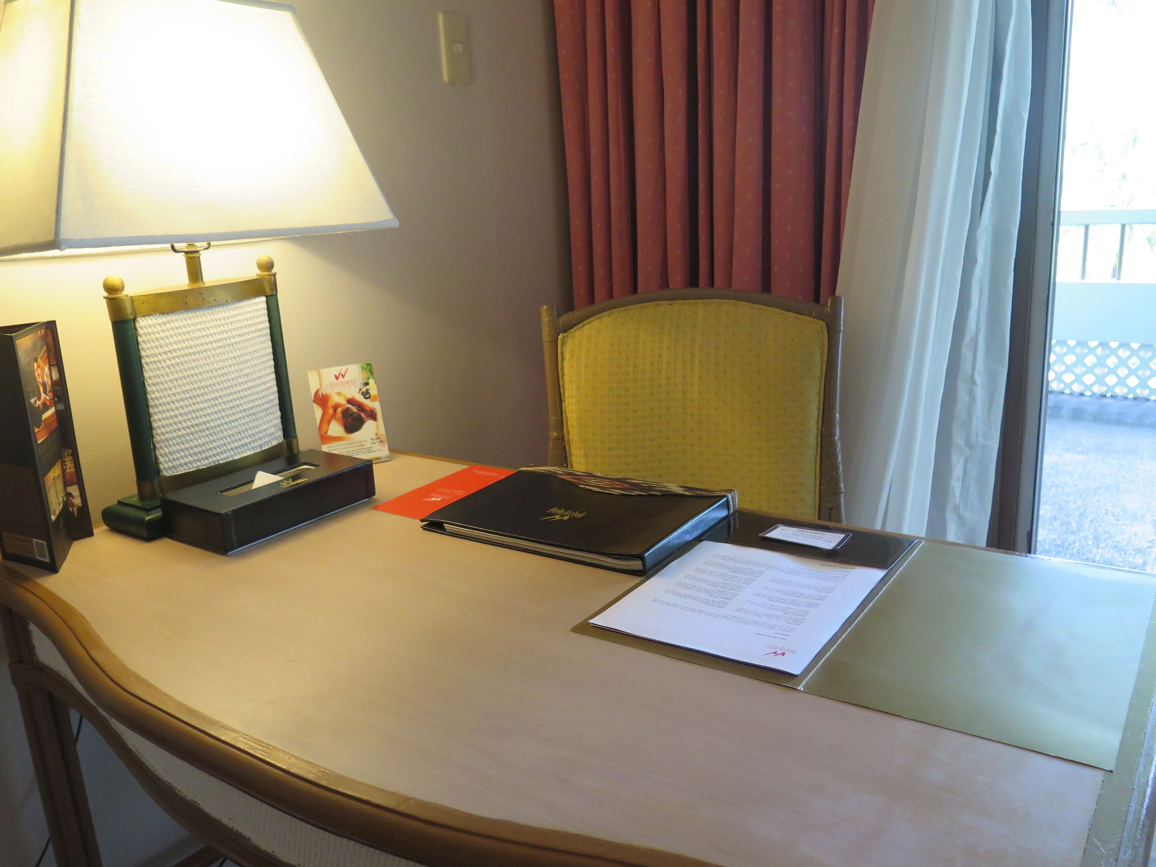 A hotel room desk with a lamp, a photo frame, a brochure, a notebook, and a window with curtains.