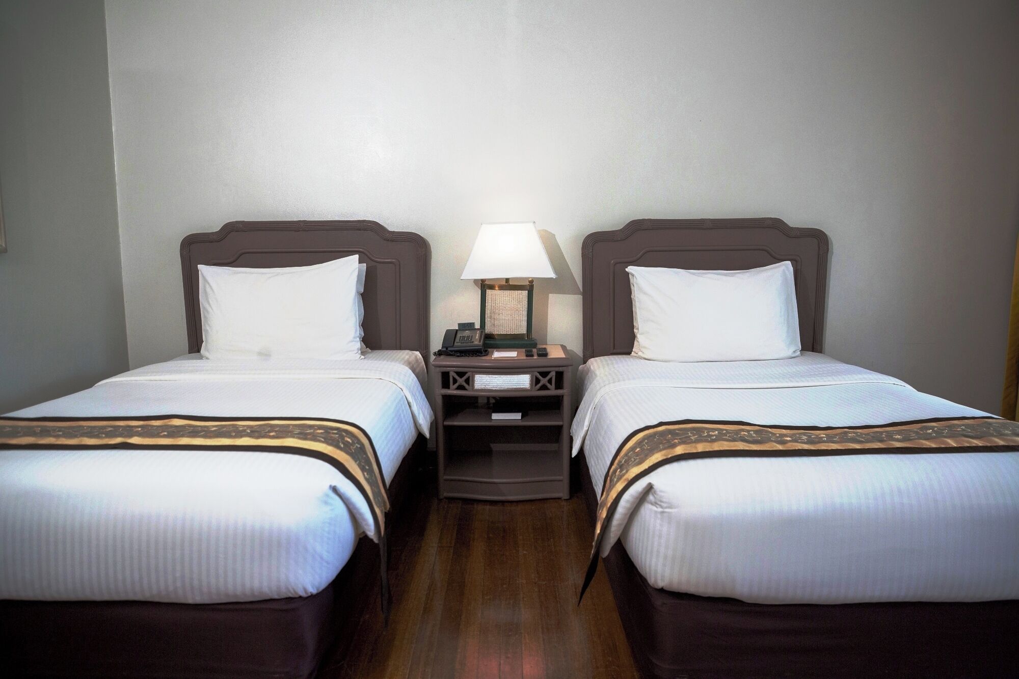 Habitación de hotel con dos camas individuales, cada una con cabecero de madera, ropa de cama blanca y una mesita de noche con lámpara.