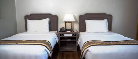 A hotel room with two single beds, each with a wooden headboard, white bedding, and a bedside table with a lamp.