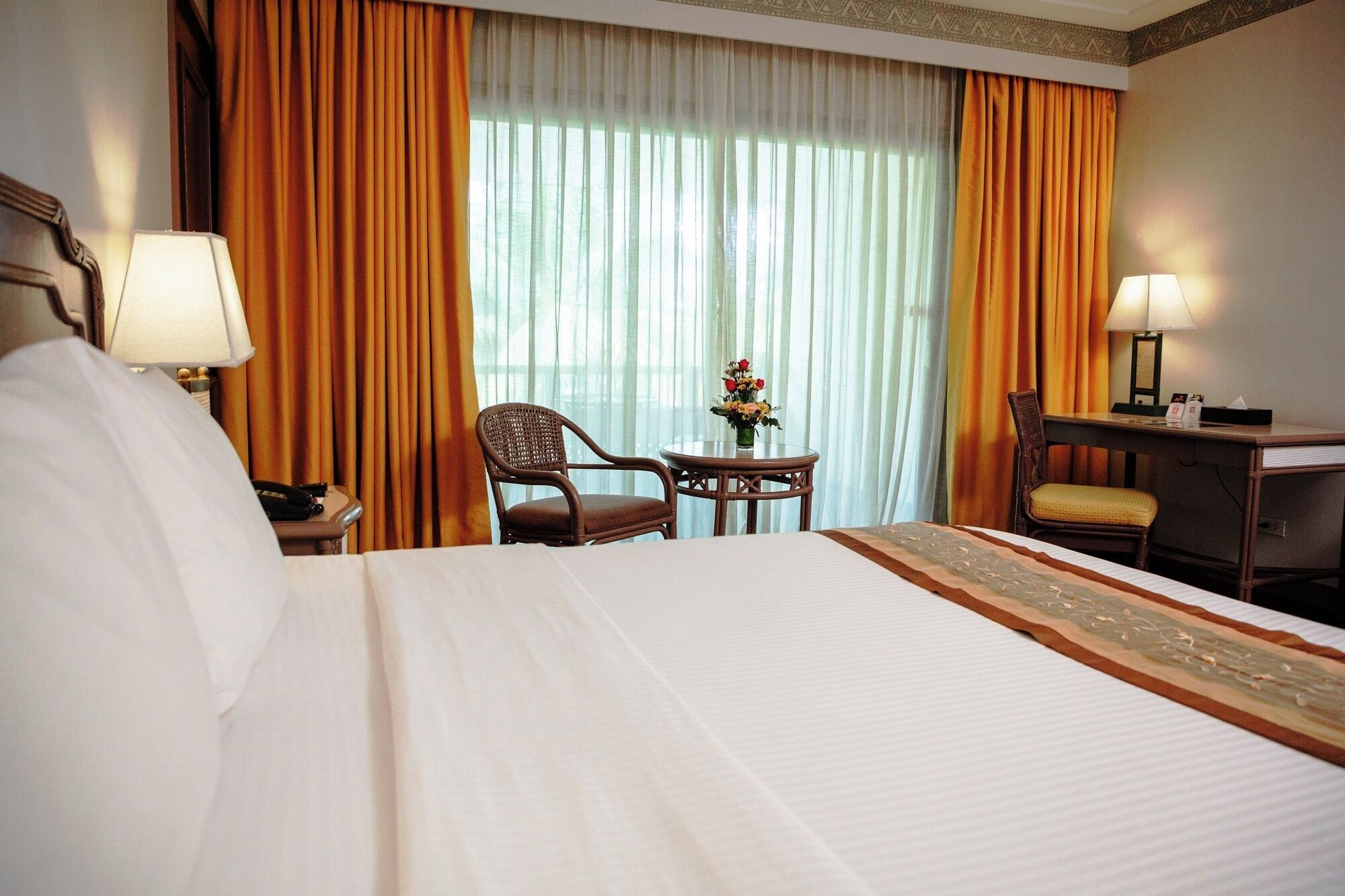 A hotel room with a bed, two chairs, a small table with a vase of flowers, and a desk with a lamp.