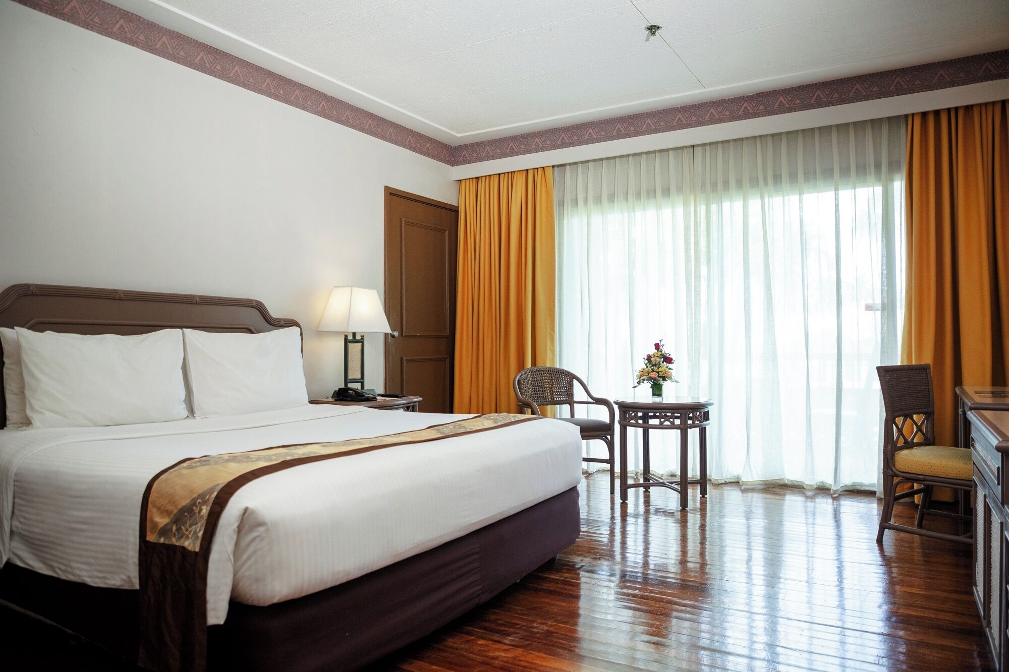 A hotel room with a large bed, a desk with a lamp, a chair, and a small table with flowers.