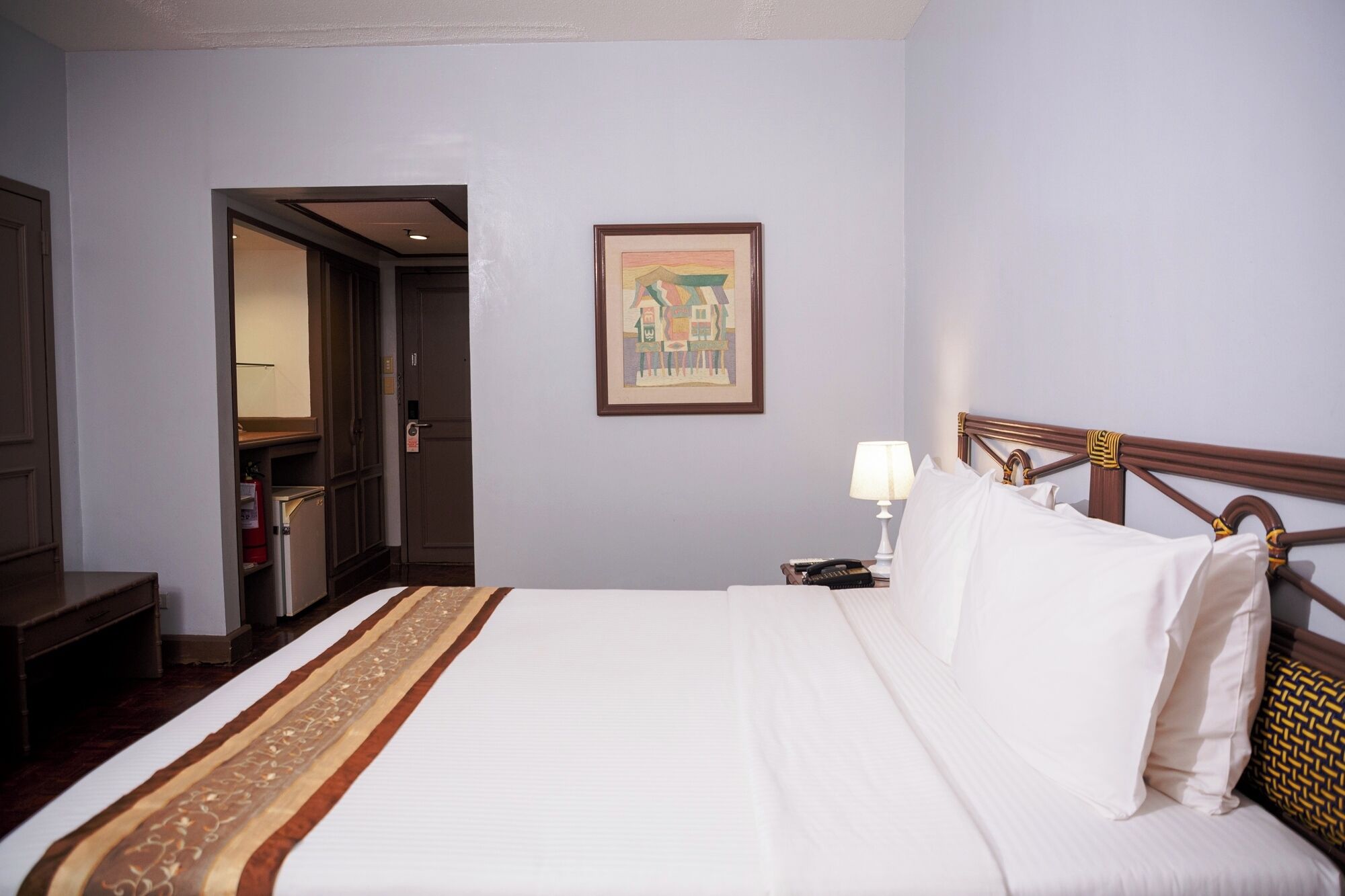 Habitación de hotel con una cama bien hecha, una lámpara de noche, una obra de arte enmarcada en la pared y una puerta que da a otra habitación.