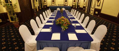 Un salón de conferencias con una larga mesa, sillas blancas y un mantel azul.
