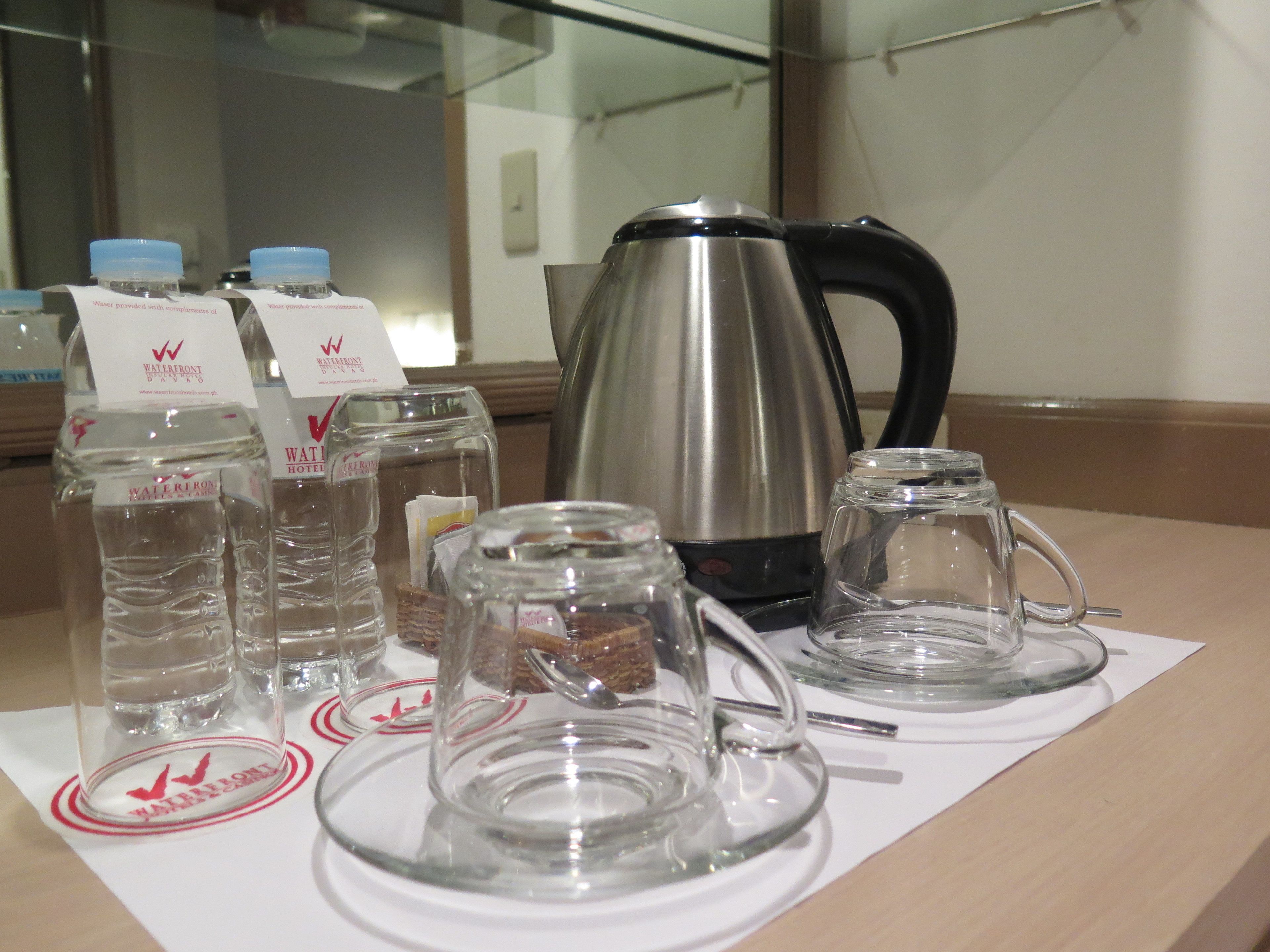 A table with water bottles, a glass carafe, a glass teapot, and a coffee pot, all on a white placemat with a logo.