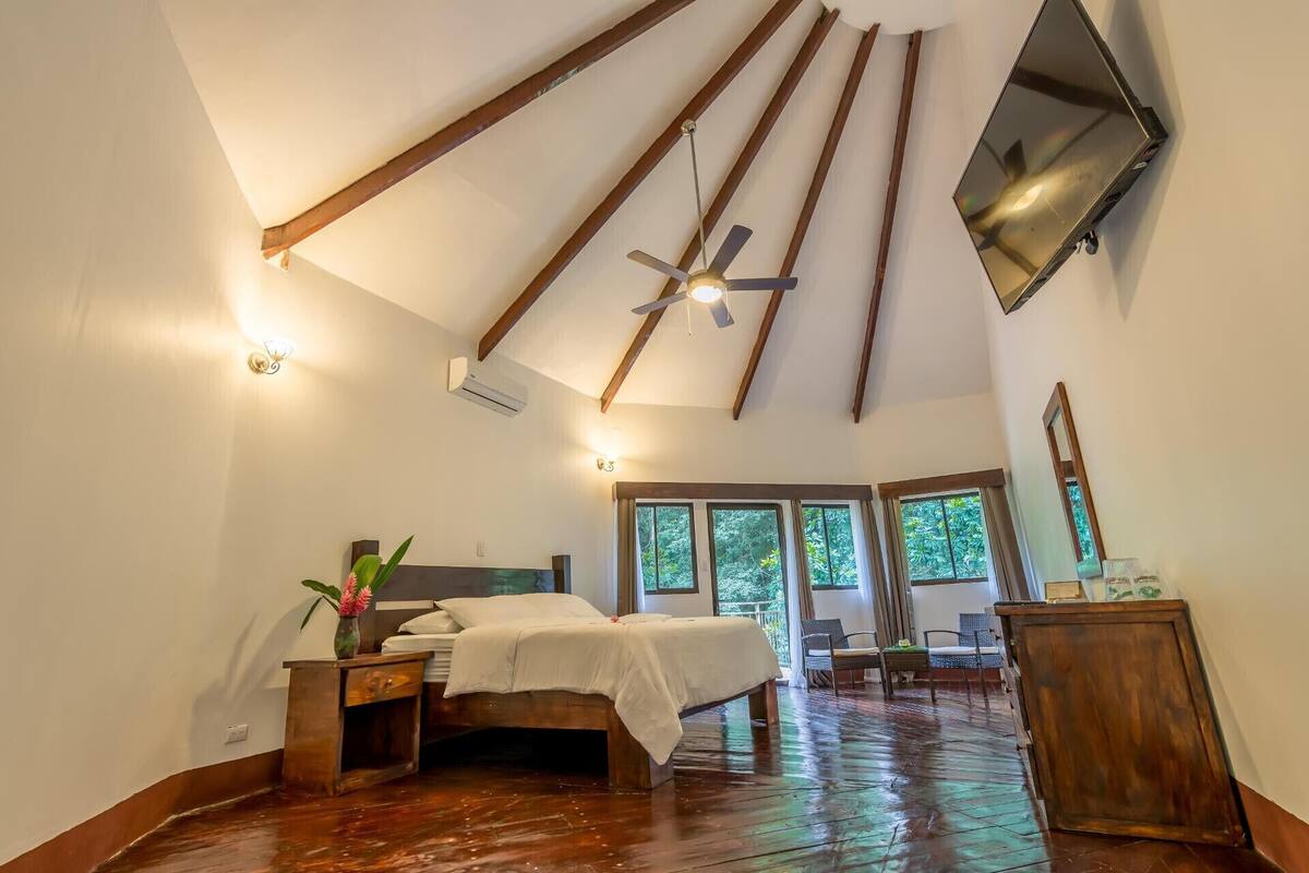 Une chambre avec un plafond en pente, des poutres en bois, un lit, des tables de chevet, un miroir et une vue sur l’extérieur grâce à de grandes fenêtres.