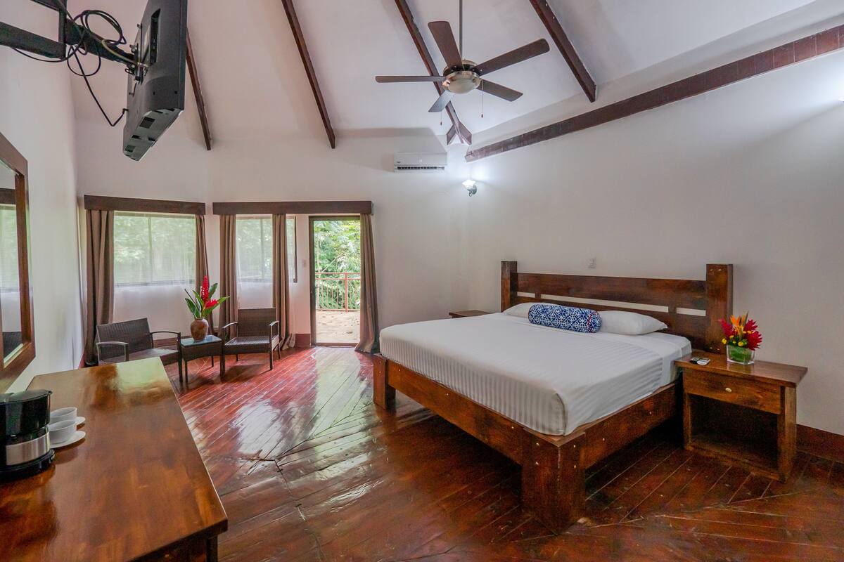 Une chambre avec un lit en bois, un bureau avec une machine à café, un ventilateur de plafond et un balcon donnant sur un paysage verdoyant.