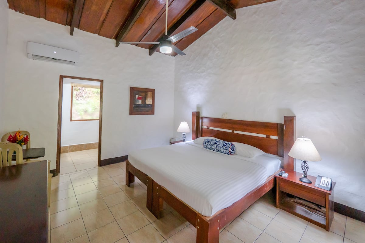 Une chambre à coucher avec un lit en bois, deux tables de chevet avec des lampes, un ventilateur de plafond et une porte donnant sur une autre pièce.
