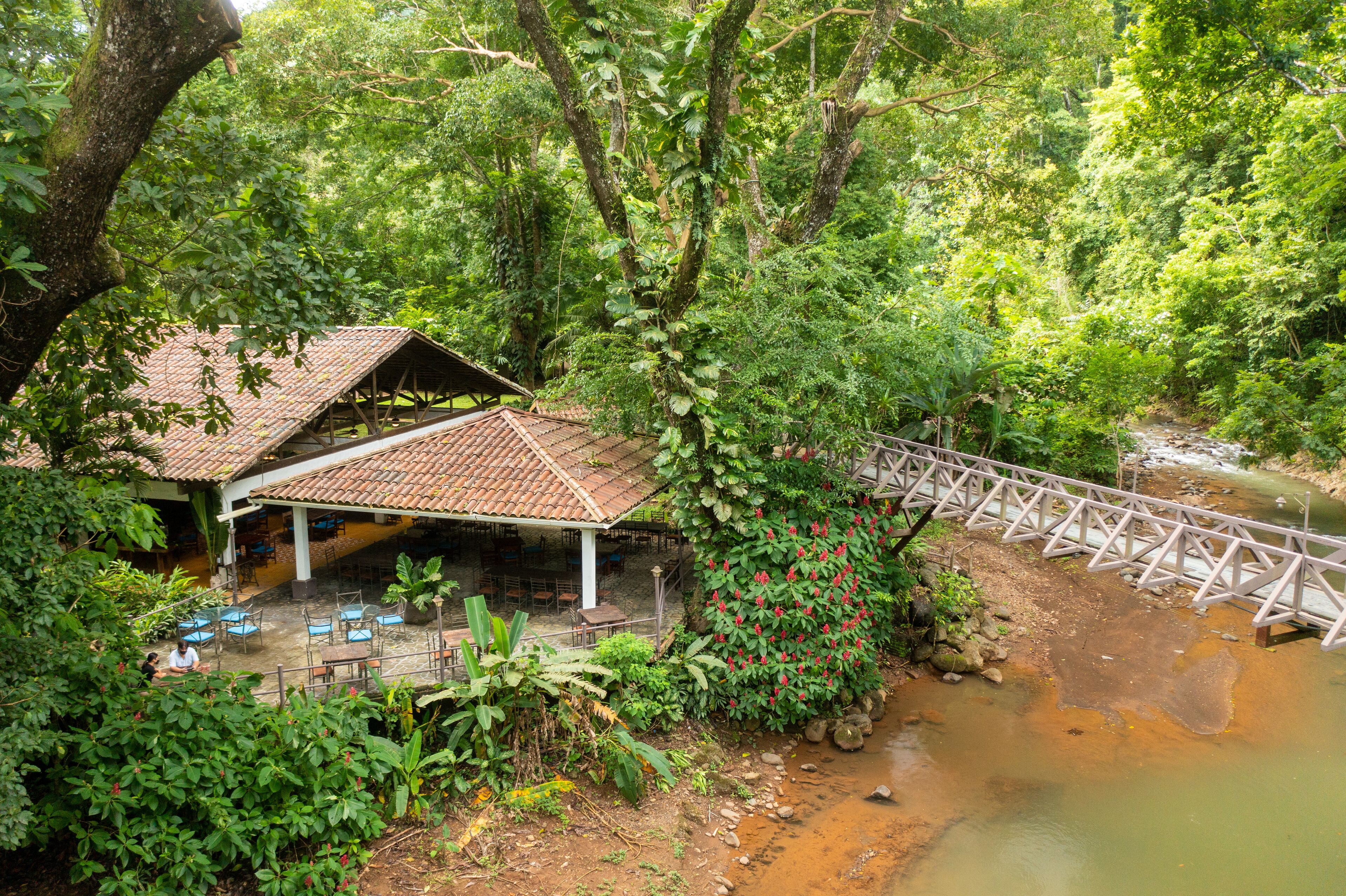 A rustic wooden structure with a tiled roof, surrounded by lush greenery and a small stream.