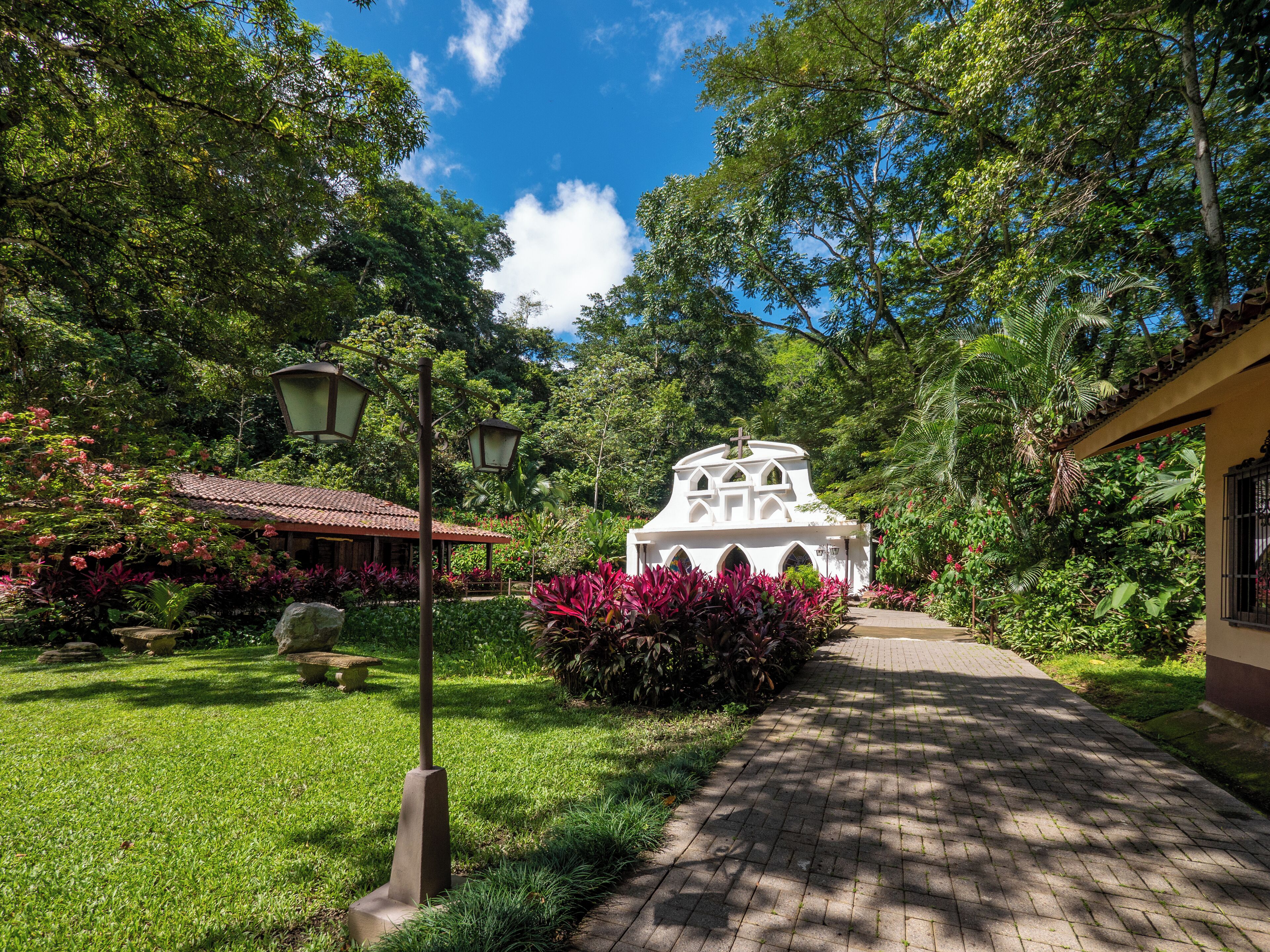 A garden pathway leading to a white building with a cross on top, surrounded by lush greenery and flowering plants.