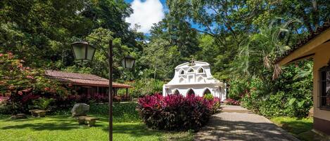 A garden pathway leading to a white building with a cross on top, surrounded by lush greenery and flowering plants.