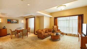 A hotel room with a living area featuring a sofa, armchair, coffee table, dining table with chairs, and a television.