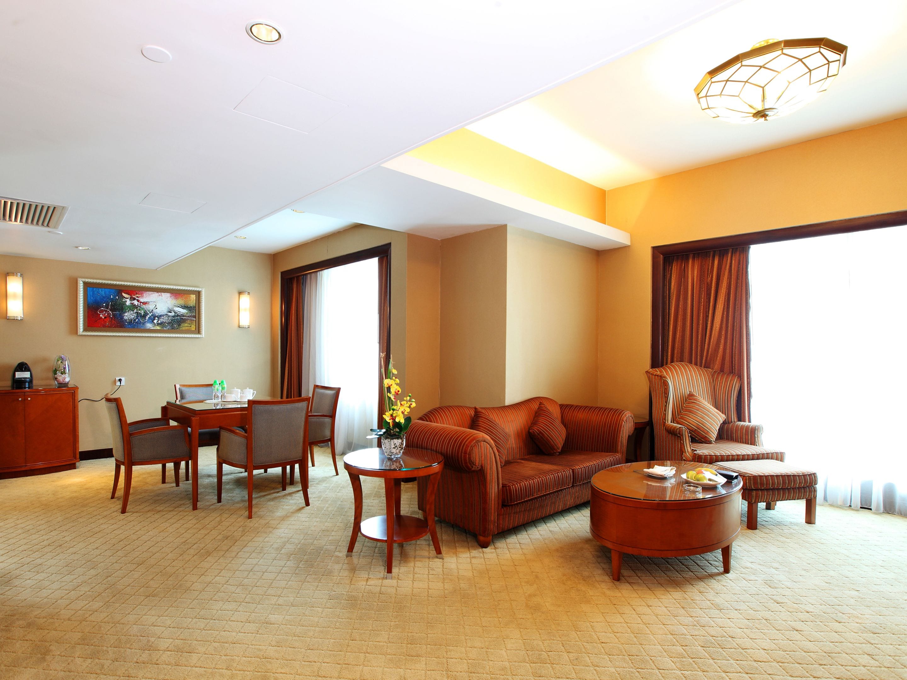 Habitación de hotel con zona de comedor, sofá, mesa de centro y un amplio ventanal con cortinas.