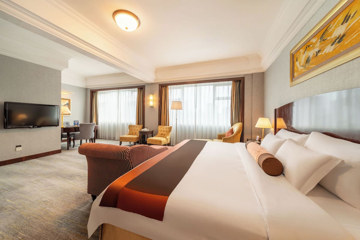 Ein Hotelzimmer mit einem großen Bett, zwei Sesseln, einem Schreibtisch, einem Flachbildfernseher und einem Bild an der Wand.