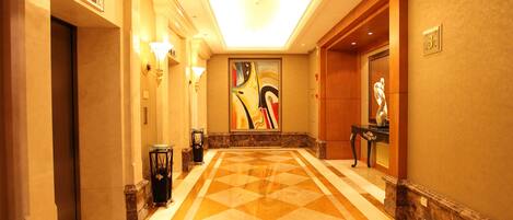 Um corredor de hotel com piso de padrão geométrico, uma grande pintura abstrata na parede e uma mesa de recepção com uma estátua.