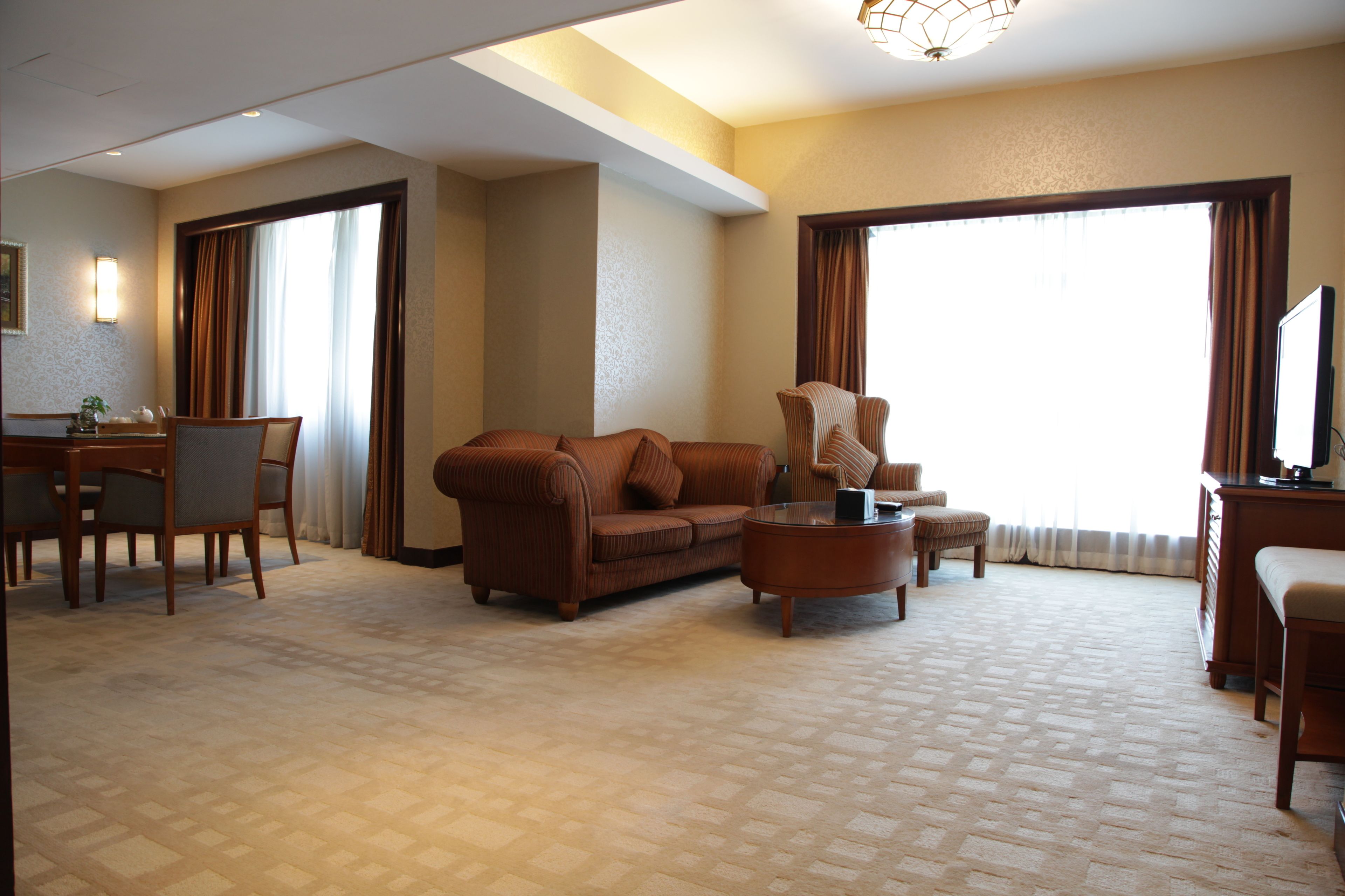 A hotel room with a sofa, a coffee table, a TV, a desk with a chair, and a large window with curtains.