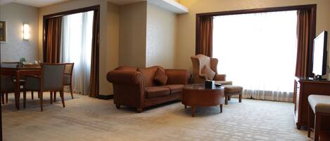 A hotel room with a sofa, a coffee table, a TV, a desk with a chair, and a large window with curtains.