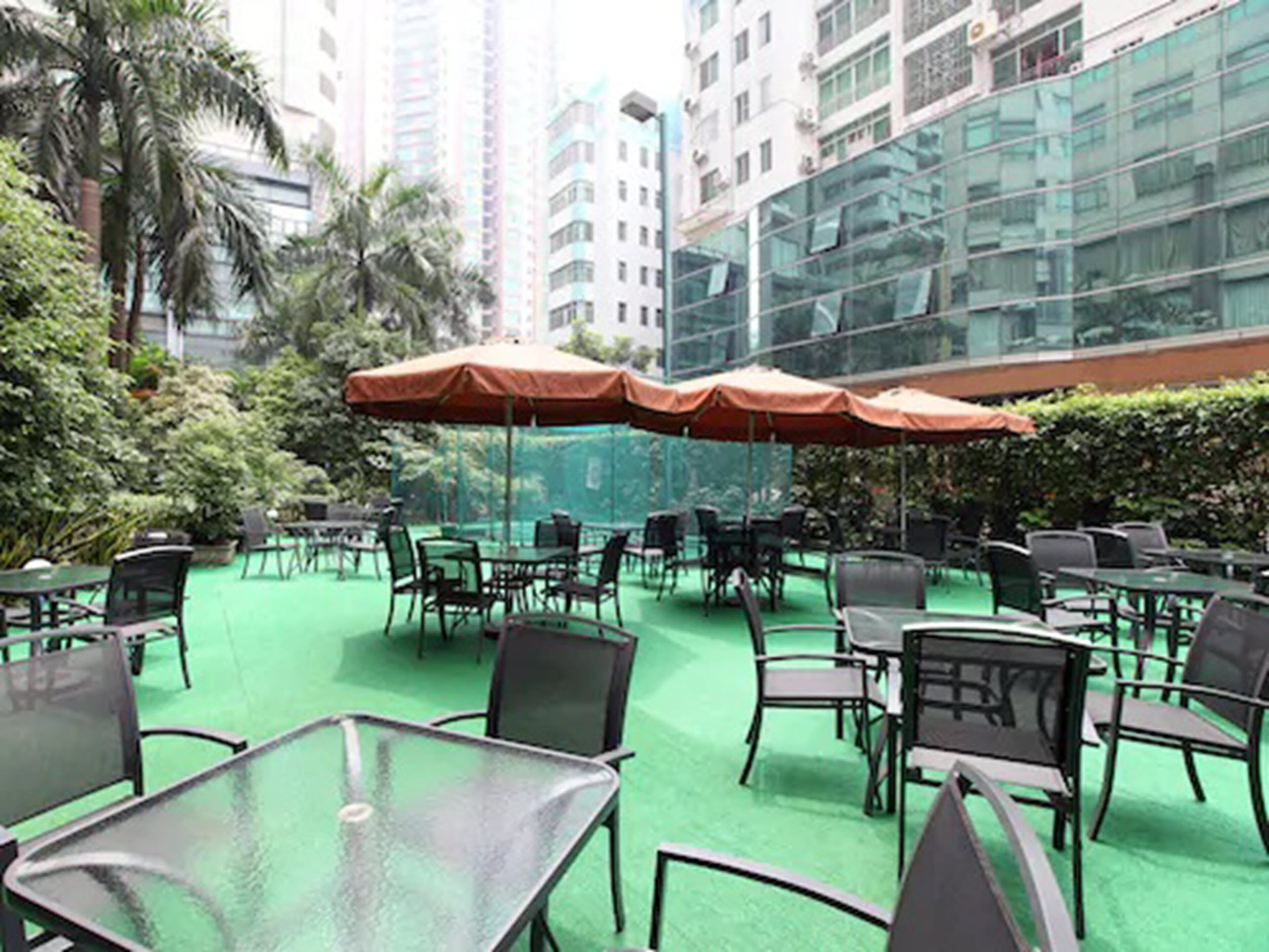 Un patio al aire libre con mesas y sillas, rodeado de follaje y edificios altos.