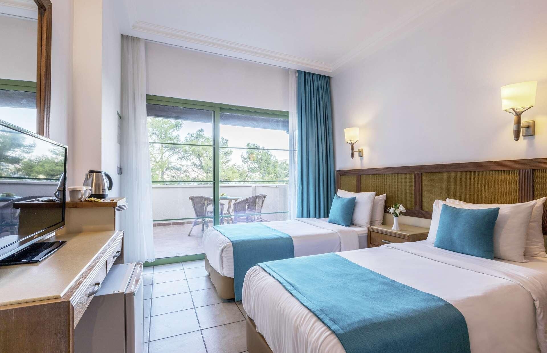 Ein Hotelzimmer mit zwei Betten, einem Schreibtisch mit Kaffeemaschine, einem Fernseher und einem Balkon mit Blick auf Bäume.