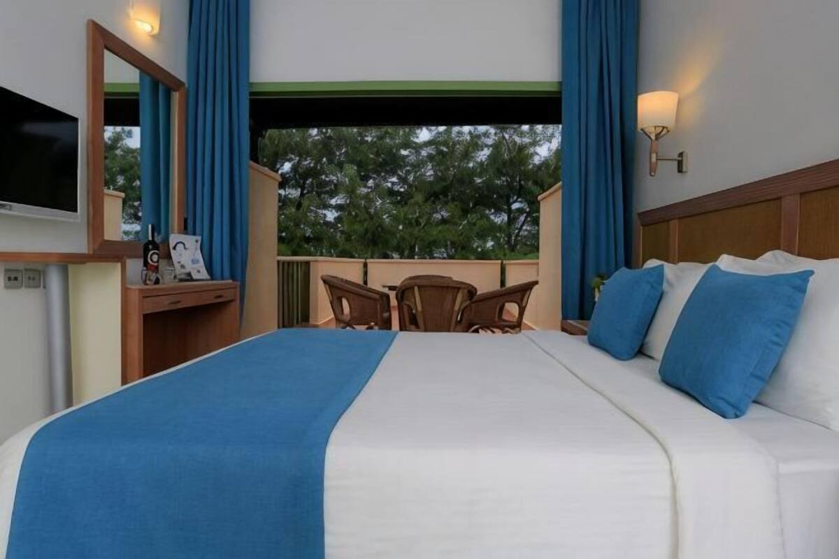 A hotel room with a large bed, a desk with a chair, a TV, and a balcony with blue curtains.