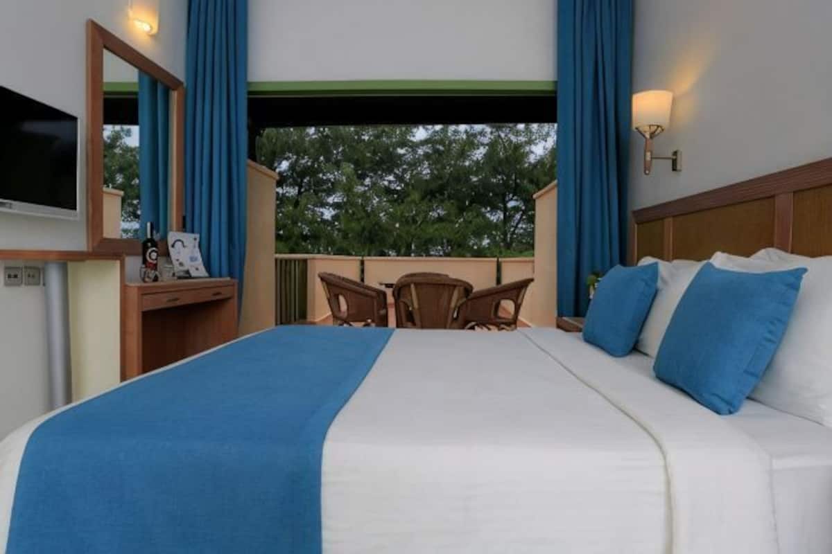 Habitación de hotel con una cama grande, un escritorio con una silla, un televisor y un balcón con cortinas azules.