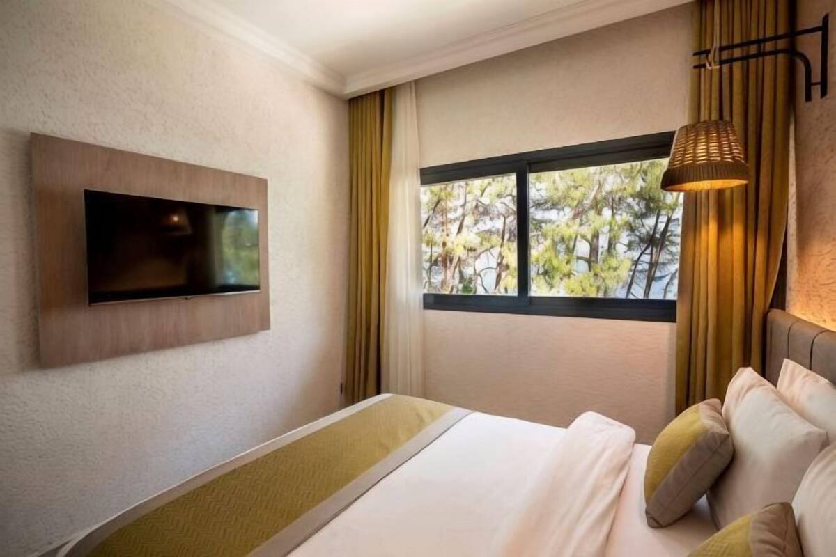 A hotel room with a bed, a TV mounted on the wall, a window with curtains, and a lamp.