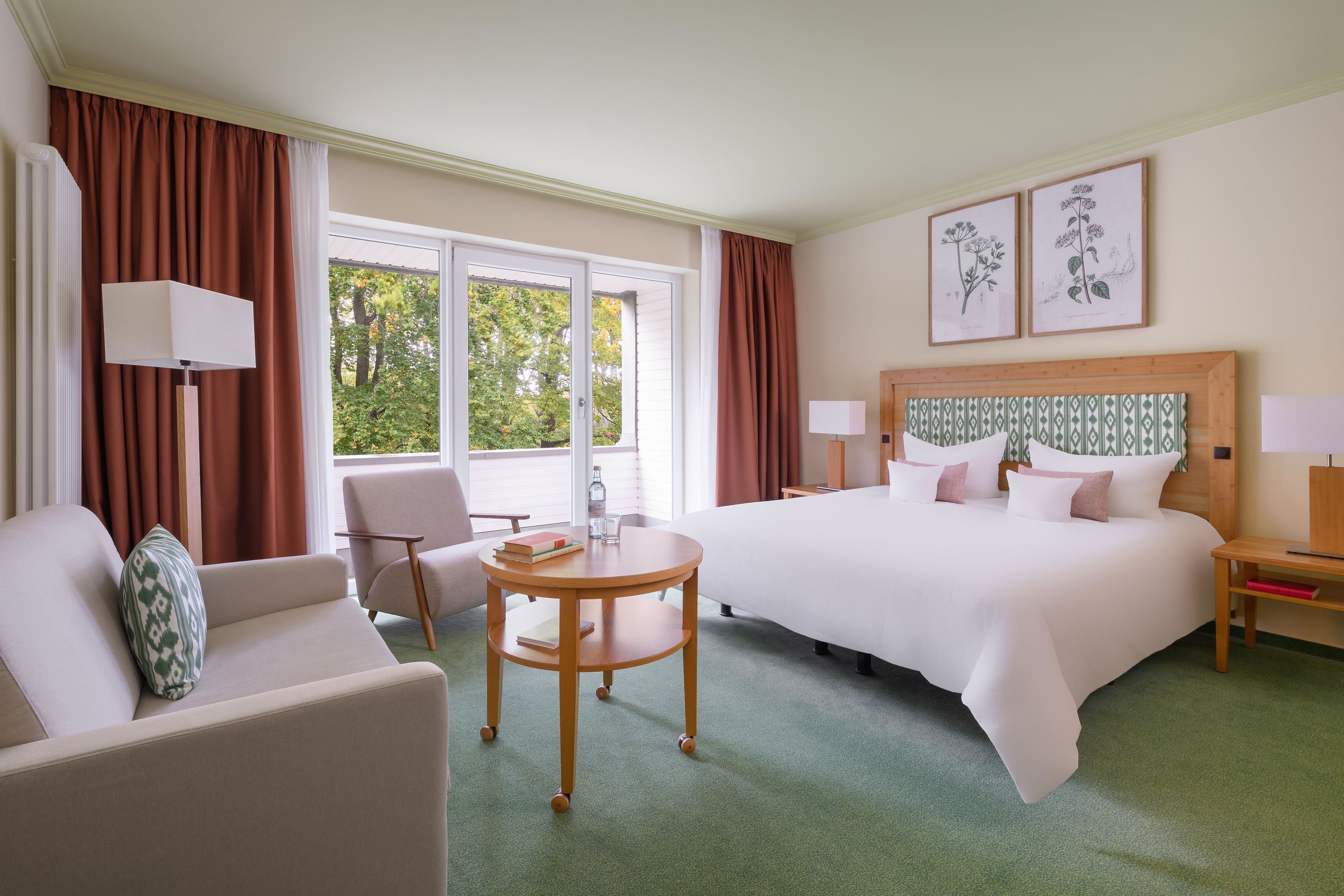 A hotel room with a large bed, a round table, a sofa, and a chair. There are two windows with curtains, a lamp on the table, and framed botanical prints on the wall.