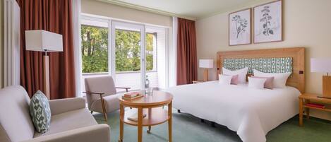 A hotel room with a large bed, a round table, a sofa, and a chair. There are two windows with curtains, a lamp on the table, and framed botanical prints on the wall.