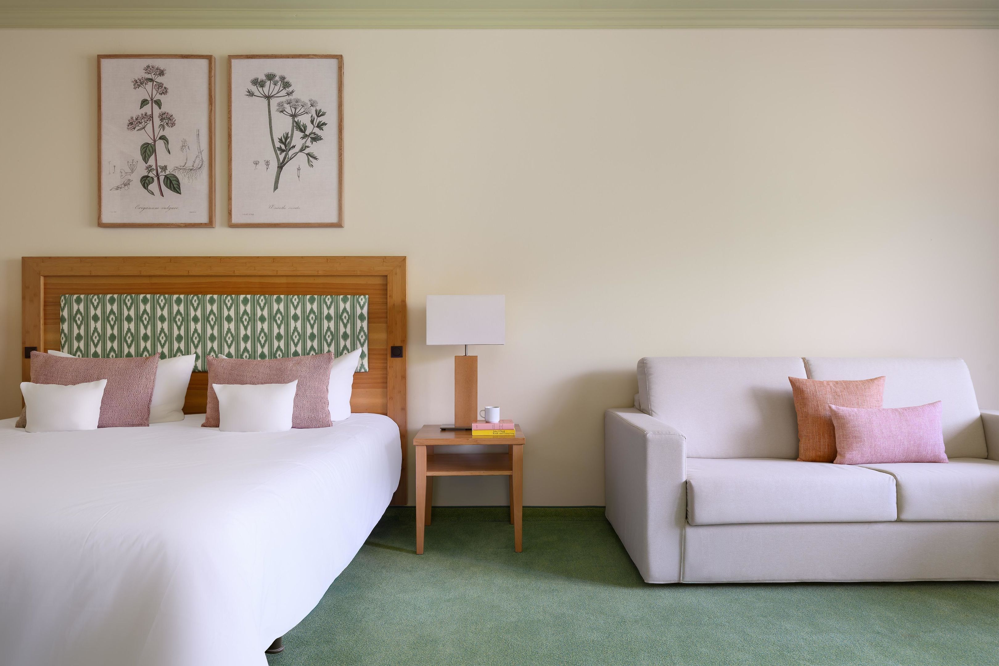 A hotel room with a bed, a nightstand, a lamp, a sofa, and two framed botanical illustrations on the wall.
